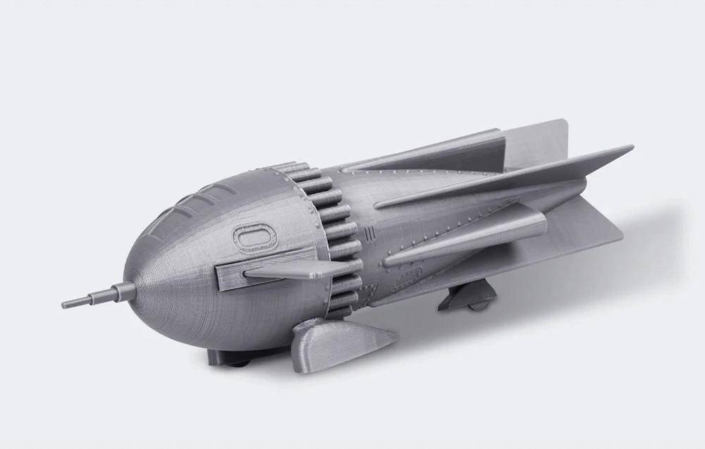 Ein 3D-gedrucktes Modell eines stilisierten Raumschiffs mit Flossen und einer abgerundeten Front, das auf einer flachen Oberfläche vor einem einfarbigen Hintergrund ruht.