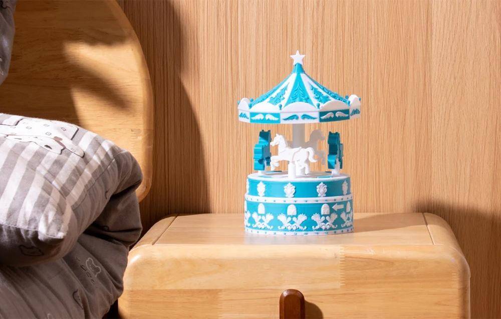 Ein Spielzeigkarussell mit Pferden in Blau und Weiß steht auf einem Holztisch im Sonnenlicht. Ein Teil einer Person, die ein graues Hemd trägt, ist teilweise sichtbar.