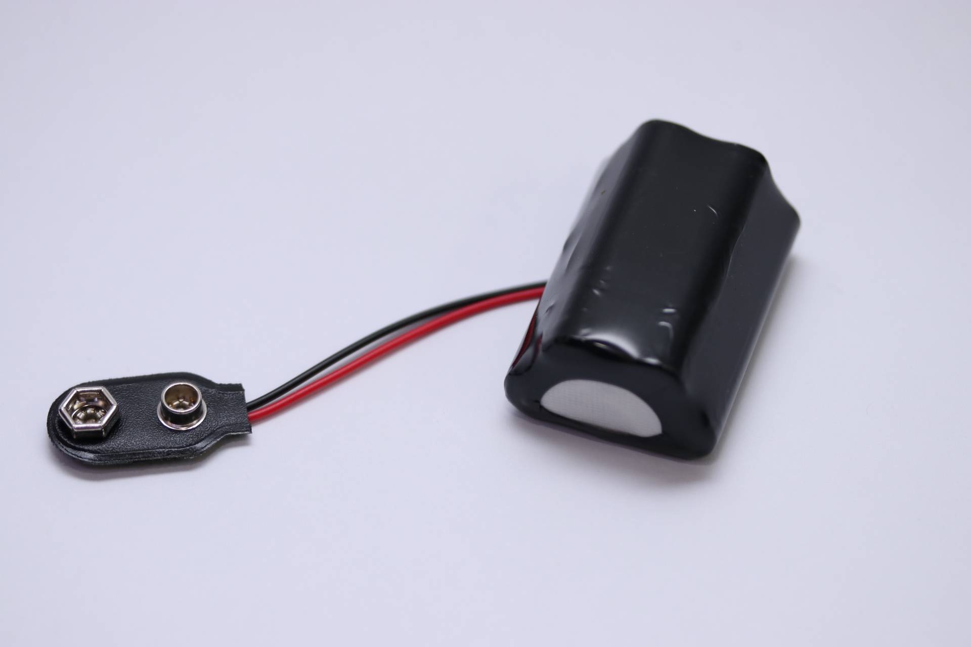 Eine schwarze, rechteckige Batterie, die in Plastik eingewickelt ist und mit roten und schwarzen Kabeln versehen, vor einem einfarbigen weißen Hintergrund positioniert.