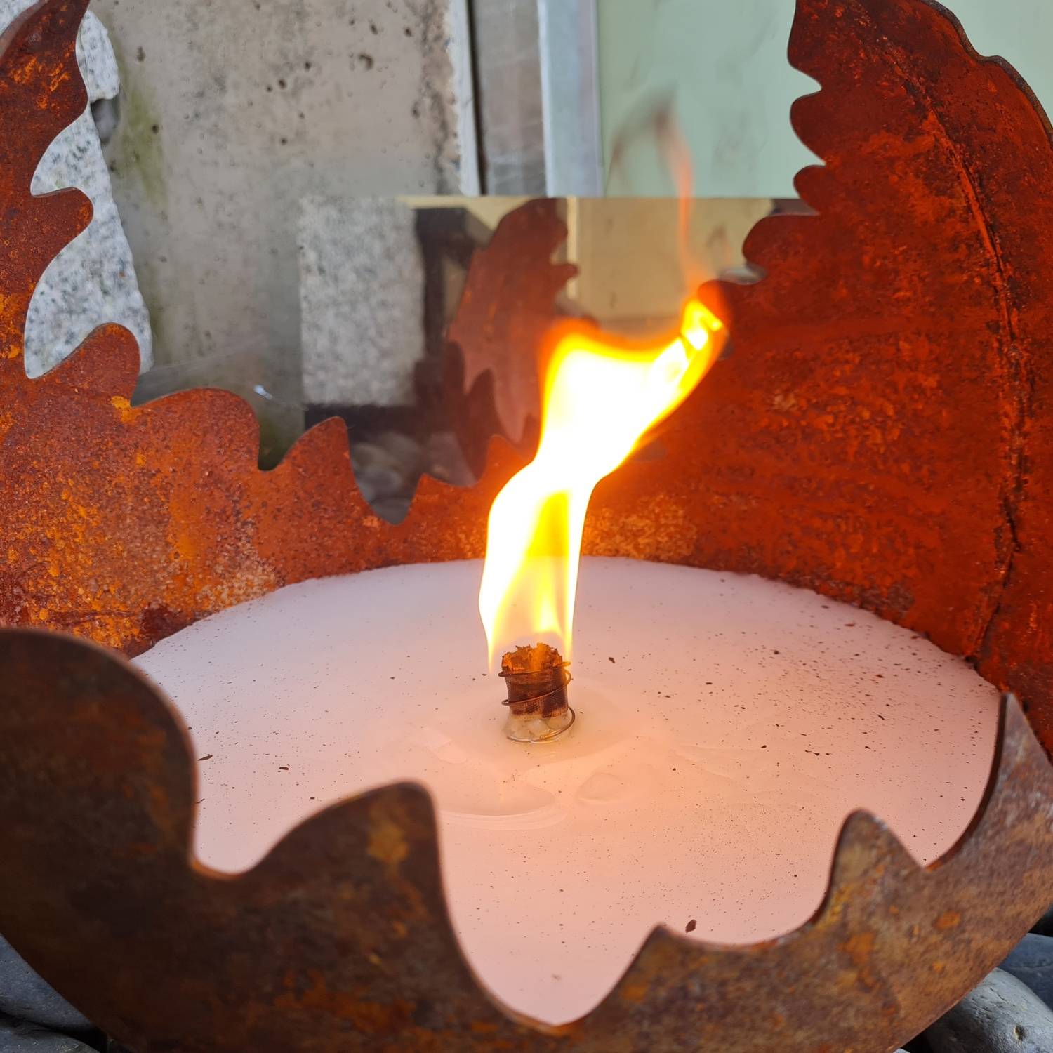 Eine große Kerze brennt mit heller Flamme in einem dekorativen Metallhalter und erzeugt eine warme Atmosphäre vor einem strukturierten Hintergrund.