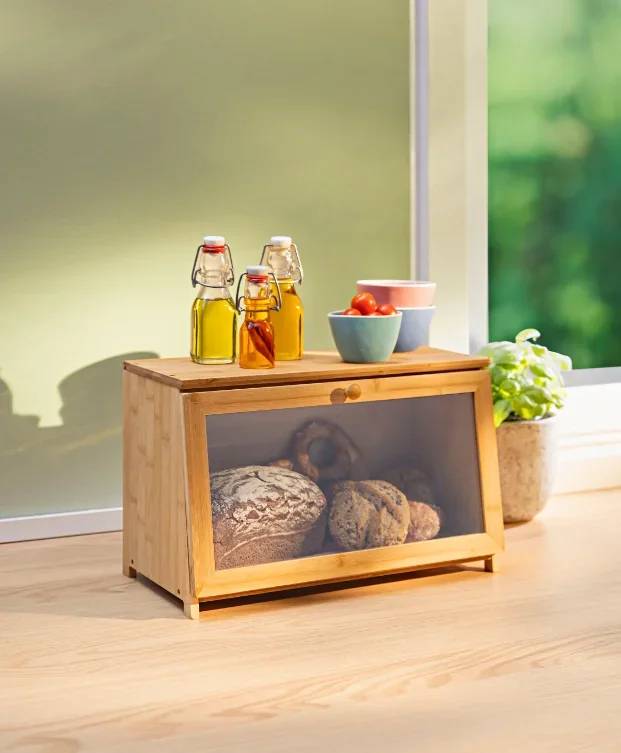 Eine Holzbrotdose mit Glasfront enthält zwei Brote. Darauf stehen Öl- und Essigflaschen, eine Tasse und eine Schüssel mit Tomaten. Sonnenlicht durchflutet den Raum.