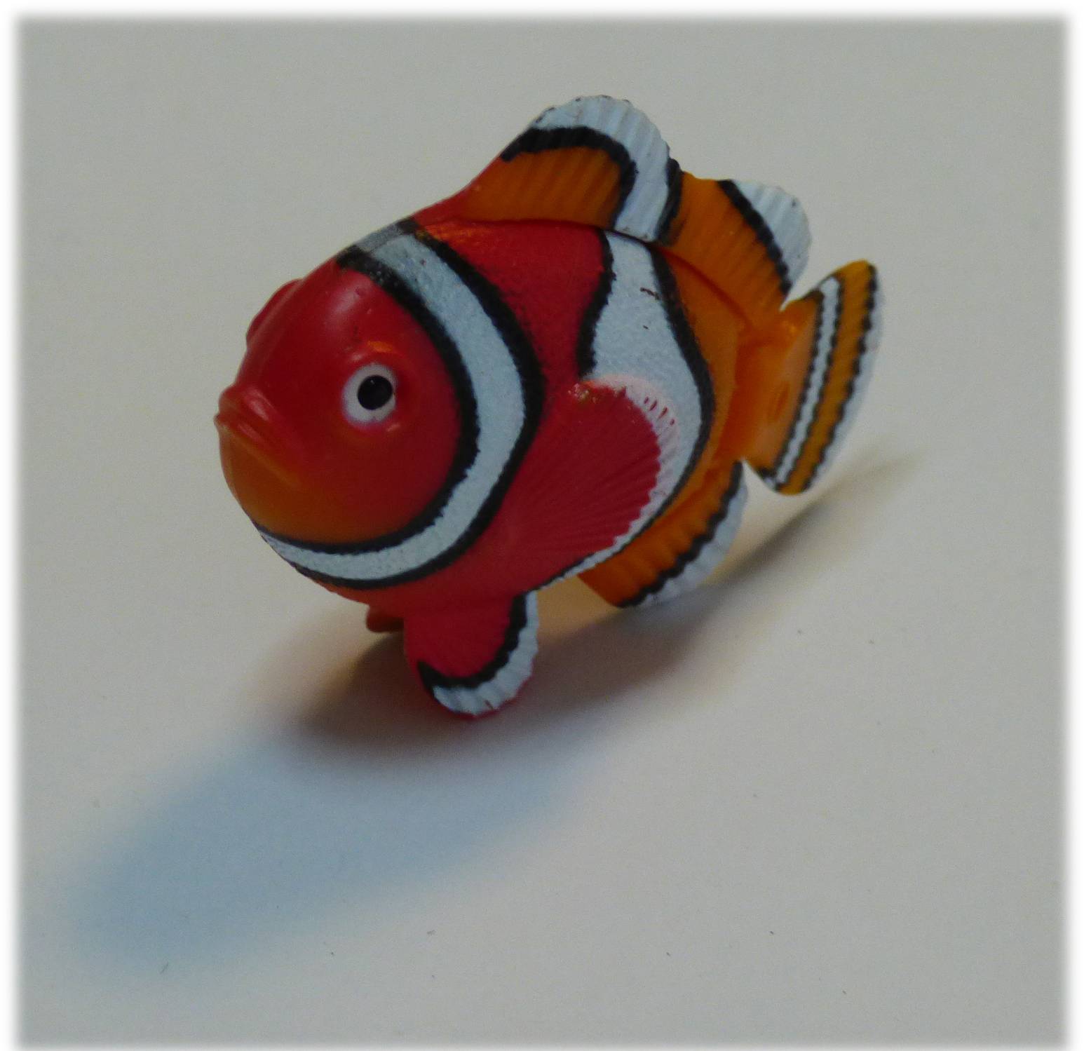 Eine kleine, bunte Clownfisch-Spielfigur mit orange-, weißen und schwarzen Streifen ist vor einem einfarbigen Hintergrund zu sehen.