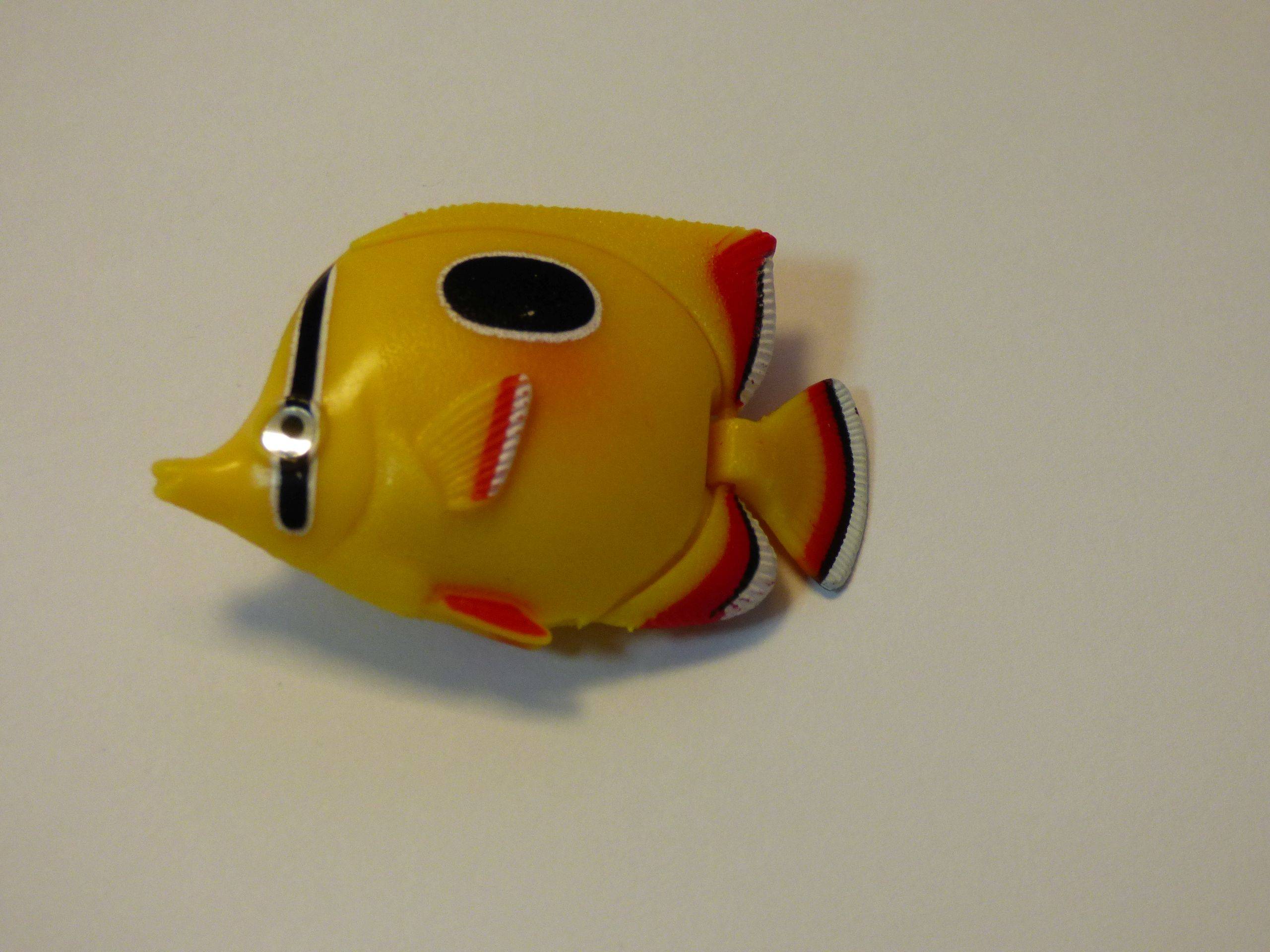 Ein kleines, hell gelbes Fisch-Spielzeug mit roten und schwarzen Akzenten, geformt wie ein tropischer Fisch, auf einer schlichten weißen Oberfläche platziert.