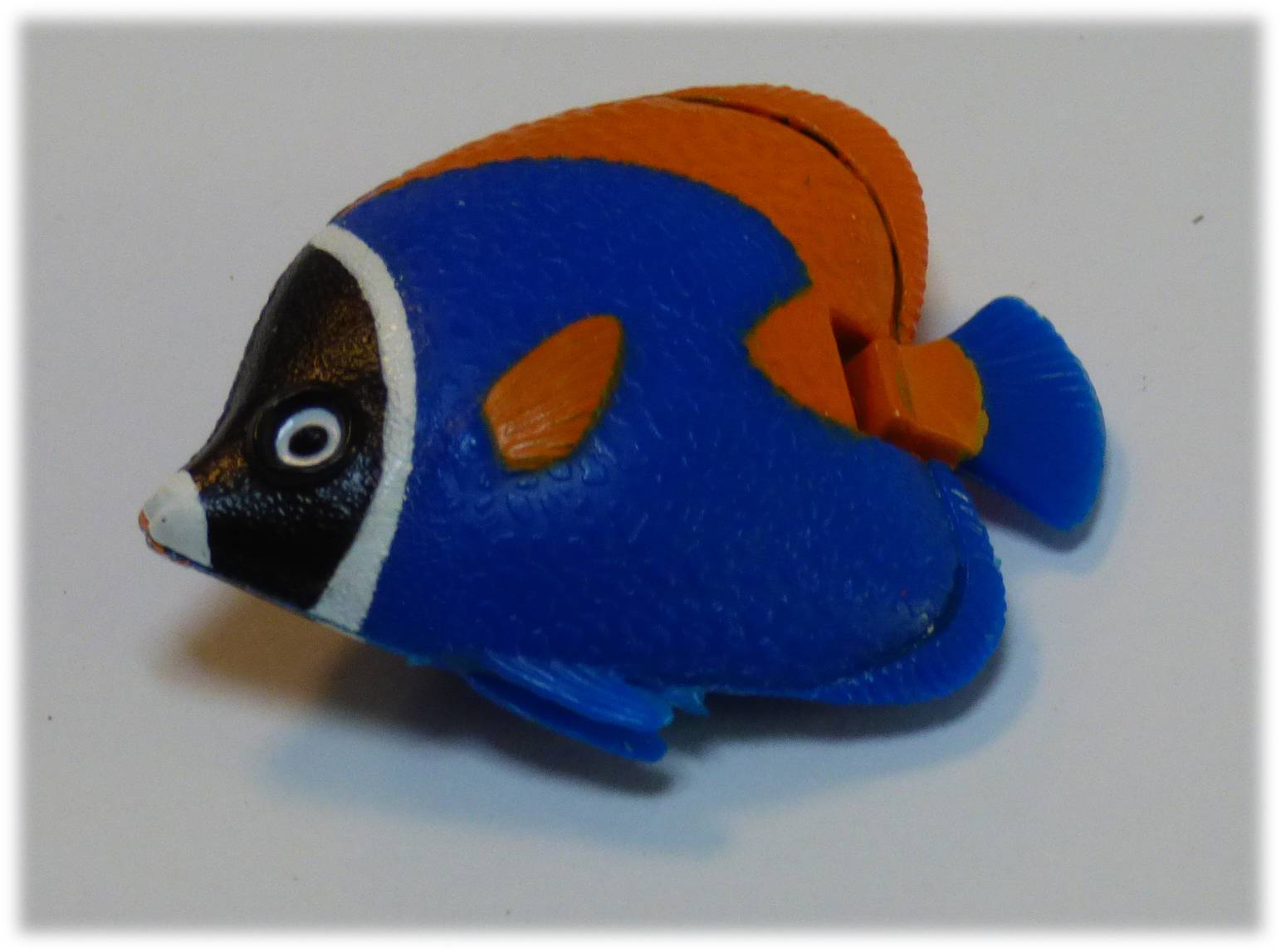 Ein kleiner blauer und oranger Spielzeugfisch mit schwarzen und weißen Markierungen, der auf einer einfarbigen Oberfläche liegt.
