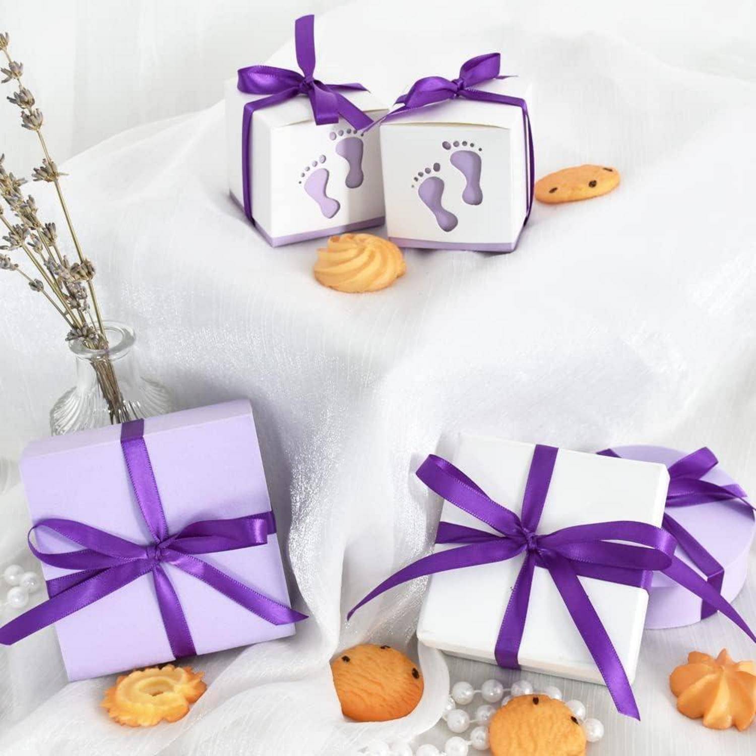 Geschenkkartons, mit lila Bändern umwickelt, umgeben von Keksen und Perlen. Eine Karte mit Babyfußabdrücken trägt die Aufschrift ‚winziges Geschenk'.