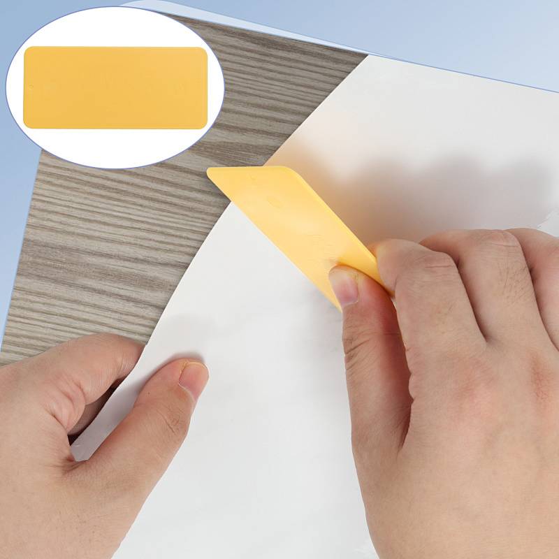 Eine Hand verwendet ein gelbes Eckwerkzeug, um ein weißes Blatt auf einer grauen Oberfläche zu glätten. Eine Einblendung zeigt eine Nahaufnahme der Kante des gelben Werkzeugs.