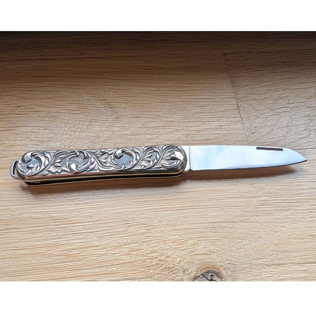 Ein kunstvoll gearbeitetes Klappmesser mit kunstvollen Gravuren am Griff liegt auf einer Holzoberfläche, teilweise geöffnet, wobei die Metallklinge sichtbar ist.
