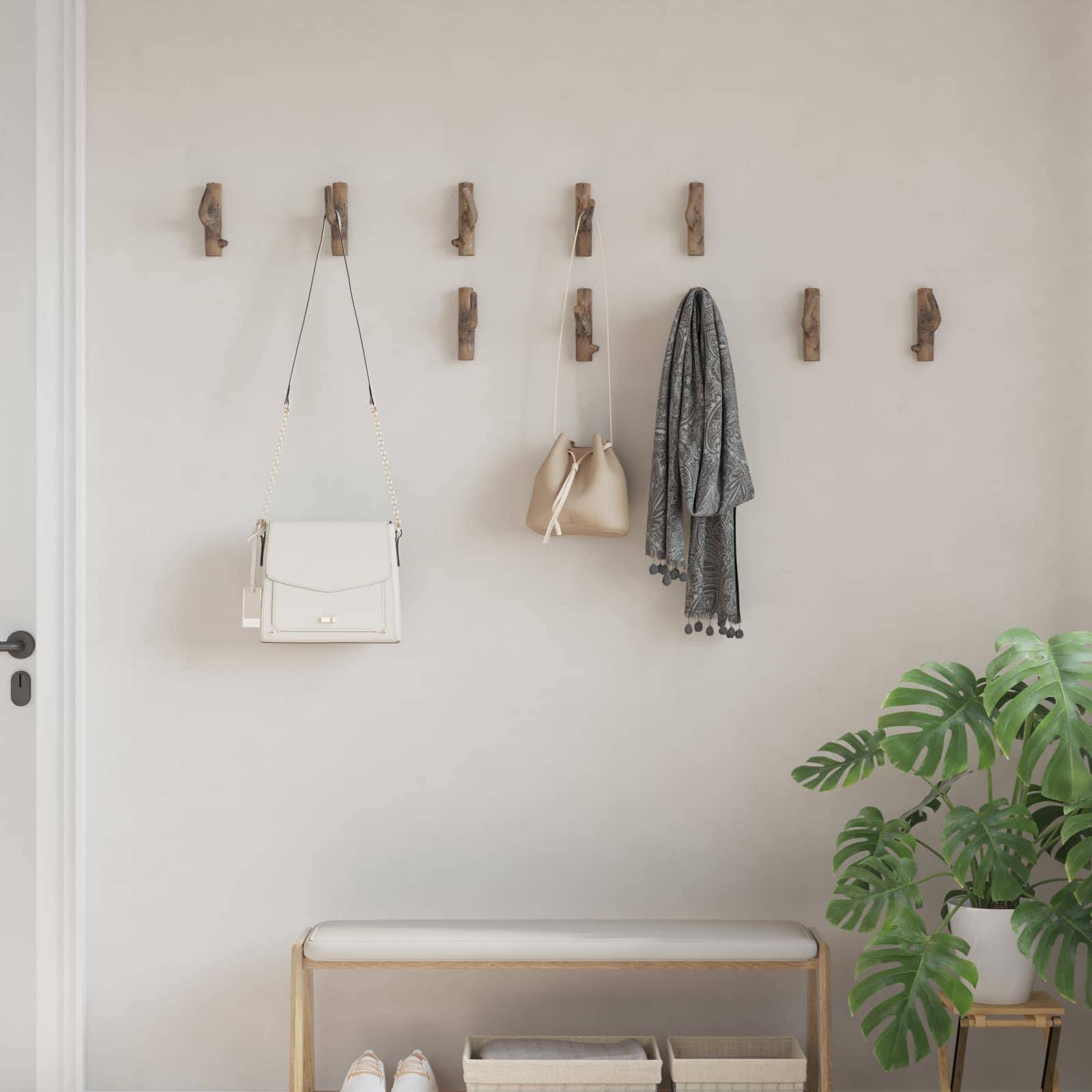 Eine minimalistische Diele mit einer Reihe von Holzhaken an der Wand, an denen eine weiße Handtasche, eine beige Tasche und ein grauer Schal hängen. Eine Bank und eine Pflanze befinden sich in der Nähe.
