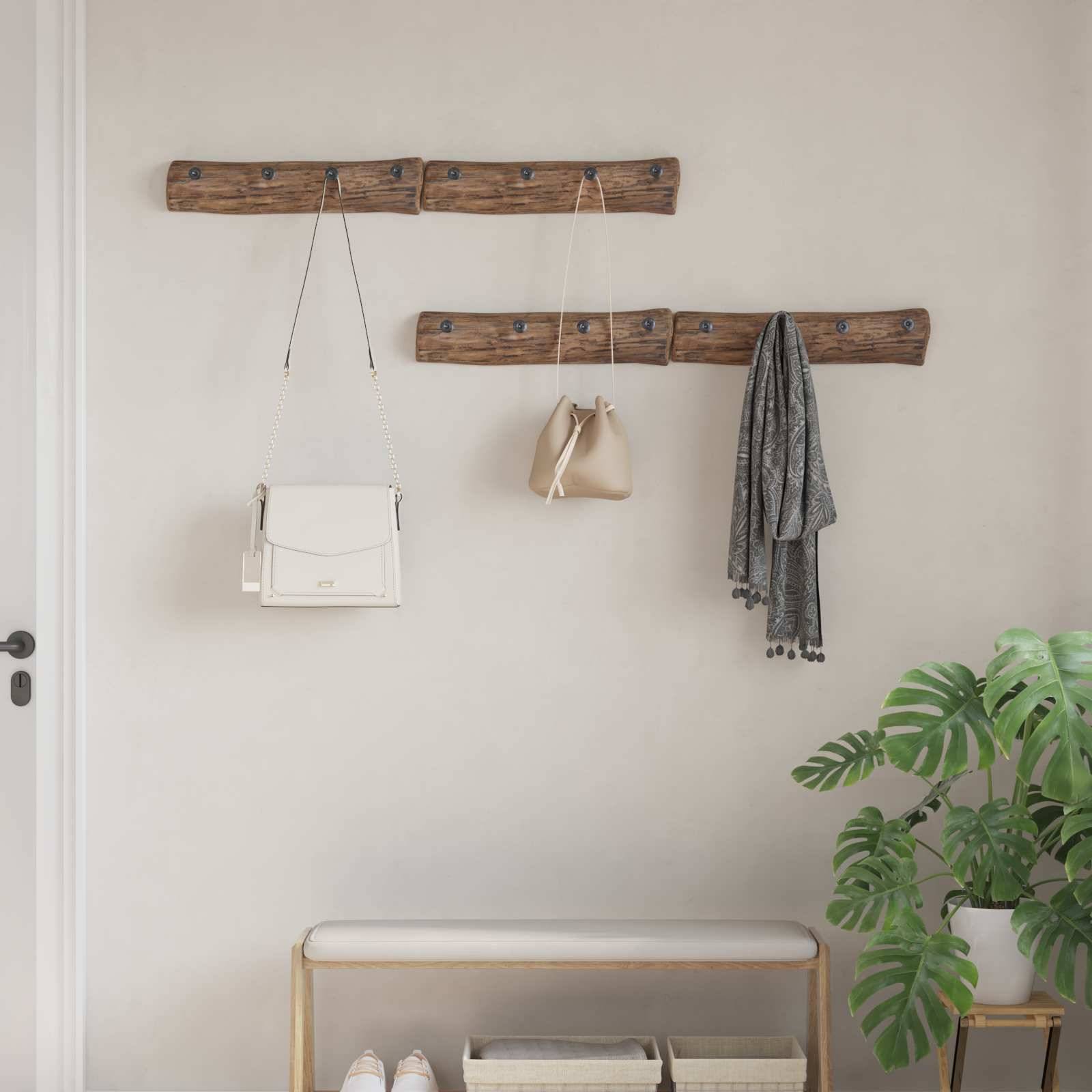 An der Wand befestigte Holzhaken halten eine weiße Handtasche, eine beige Tasche und einen grauen Schal. Darunter steht eine Bank neben einer Topfpflanze, vor einer hellen Wand.