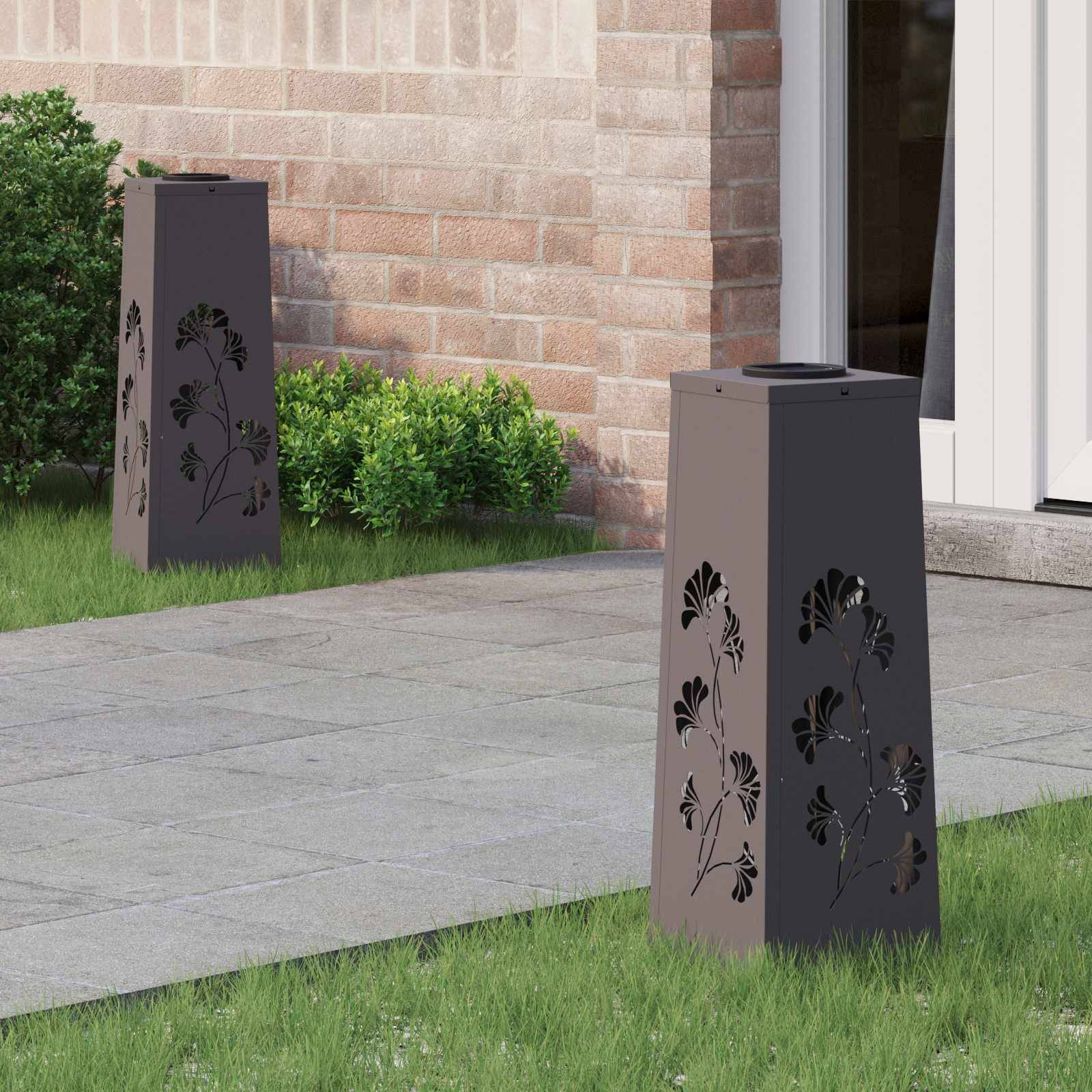 Eine Gartenszene mit zwei dekorativen Metalllaternen mit Blattausschnitten, die auf einer gefliesten Terrasse in der Nähe von grünen Büschen und einer Backsteinmauer platziert sind.