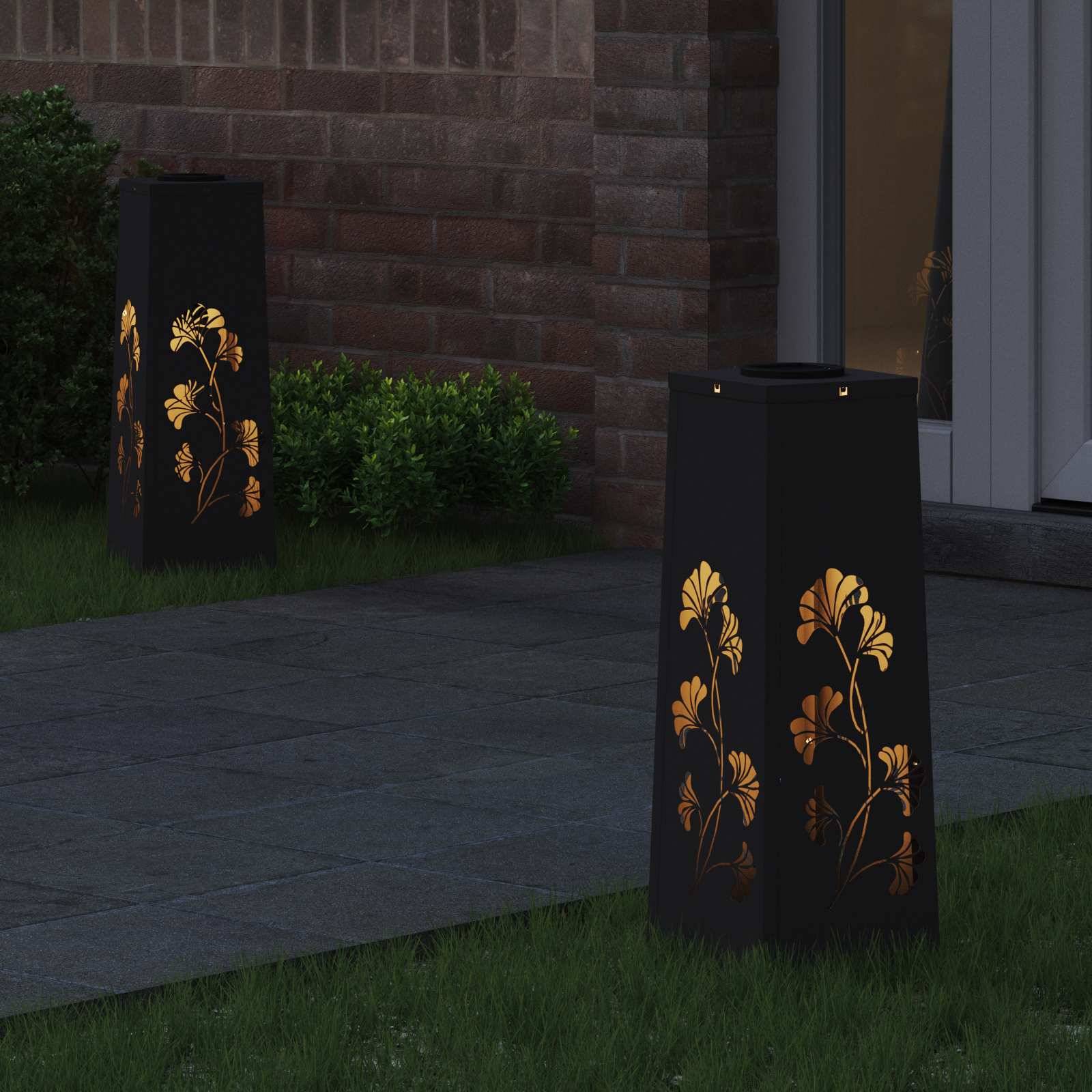 Dekorative Laternen mit Blumenausschnitten, die nachts beleuchtet sind, platziert auf einem gepflasterten Weg in der Nähe üppiger Vegetation und vor einer Ziegelwand im Hintergrund.