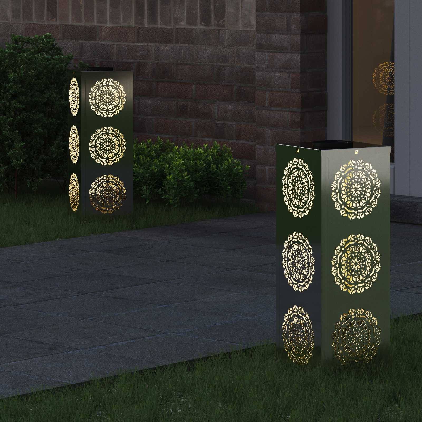 Dekorative Laternen mit aufwendigen Ausschnittmustern beleuchten nachts einen Gartenweg und werfen warmes Licht auf das umliegende Grün.