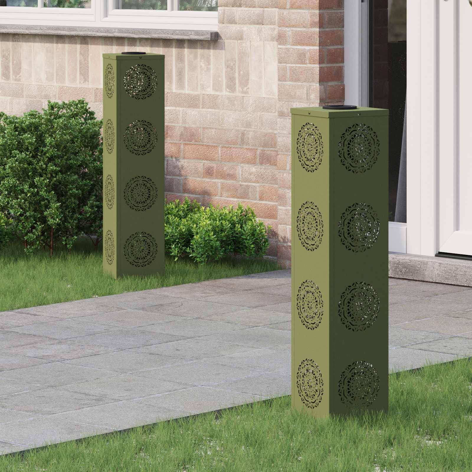 Zwei dekorative grüne Metallpfosten mit kunstvollen Mustern flankieren einen Weg, der zu einer weißen Tür führt. Eine Ziegelwand und Büsche befinden sich im Hintergrund.