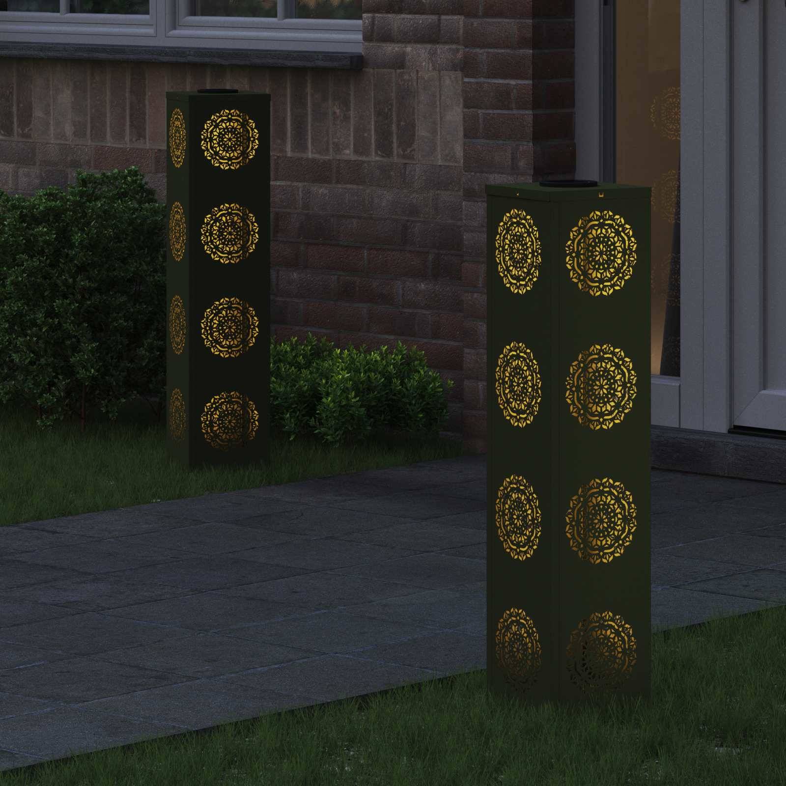 Zwei dekorative Außenlampen mit kunstvollen goldenen Mustern beleuchten einen Weg neben einem Backsteingebäude mit Grünanlage in der Nähe.