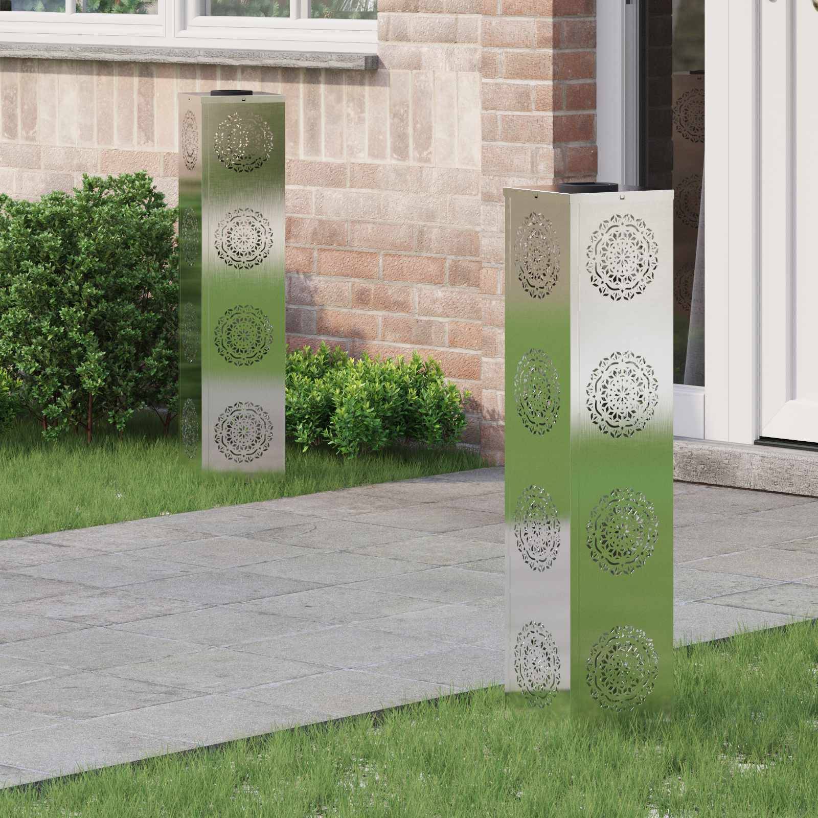 Dekorative Metall-Gartenleuchten mit verschlungenen Mustern stehen auf einem gepflasterten Weg neben einem Haus, umgeben von Gras und Sträuchern.