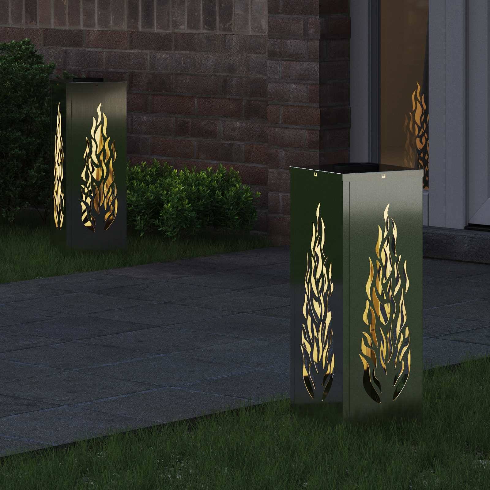 Zwei moderne Außenleuchten mit ausgeschnittenen Flammenmotiven beleuchten eine dunkle Ziegelsteinterrasse neben einer Glastür und üppigem Grün.
