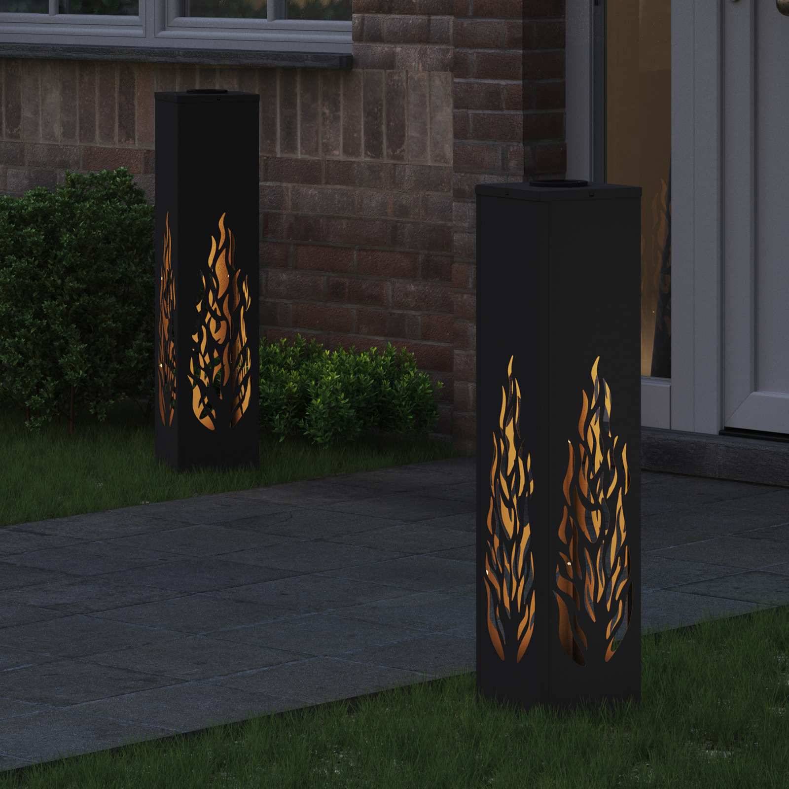 Zwei hohe Außenleuchten mit Flammenmuster beleuchten einen Weg neben einer Ziegelmauer und verleihen der Gartenumgebung eine warme Atmosphäre.