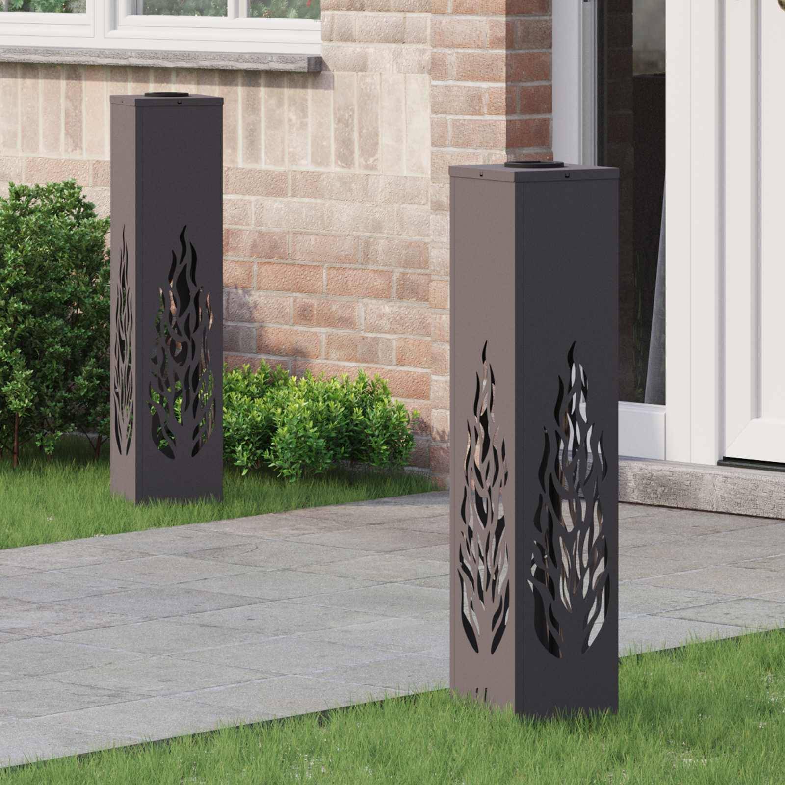 Zwei hohe, dunkel gefärbte Außenlampen mit flammenähnlichen Ausschnitten stehen neben einer Backsteinmauer auf einer Terrasse mit grünem Rasen.