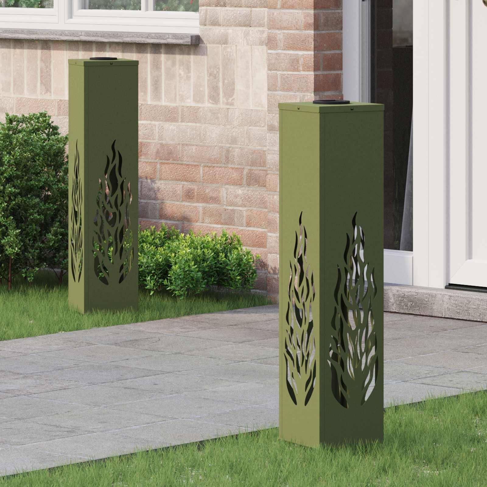 Zwei dekorative grüne Metallpfeiler mit ausgeschnittenen Flammendesigns stehen auf einem Rasen neben einer Ziegelwand und einer weißen Tür. In der Nähe befinden sich kleine Sträucher.