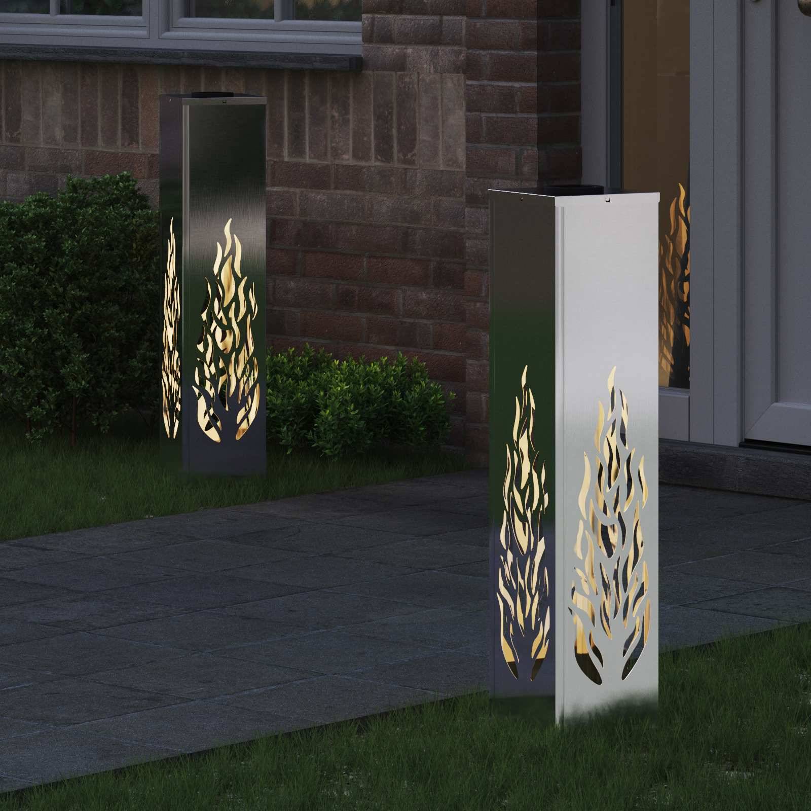 Metallische Gartenlampen mit Flammenschnittmustern beleuchten einen gepflasterten Gehweg neben einer Ziegelmauer und Grünpflanzen bei Einbruch der Dämmerung.