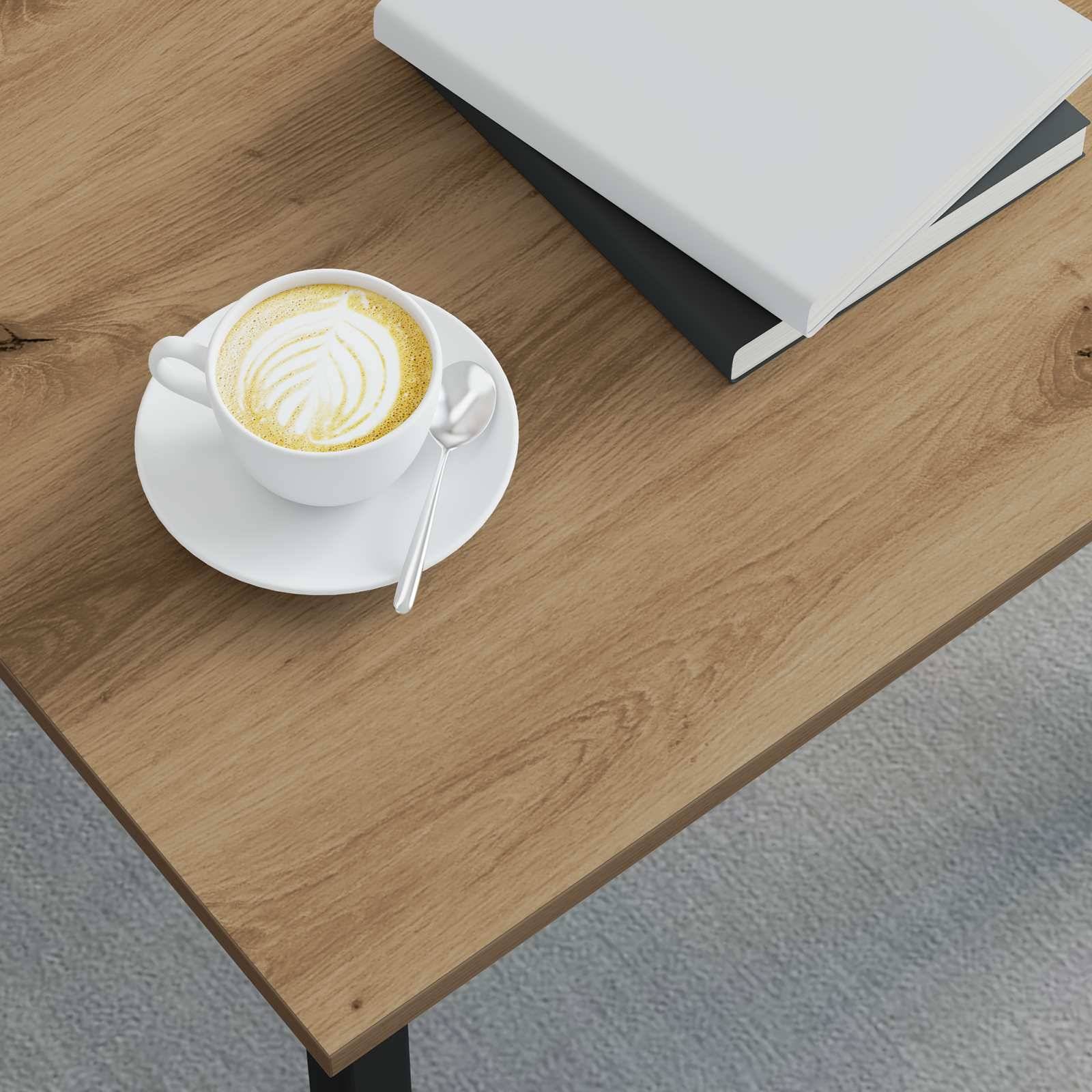 Eine Latte in einer weißen Tasse mit einem Blattmuster und einem Löffel auf einem Holztisch neben zwei geschlossenen Notizbüchern.