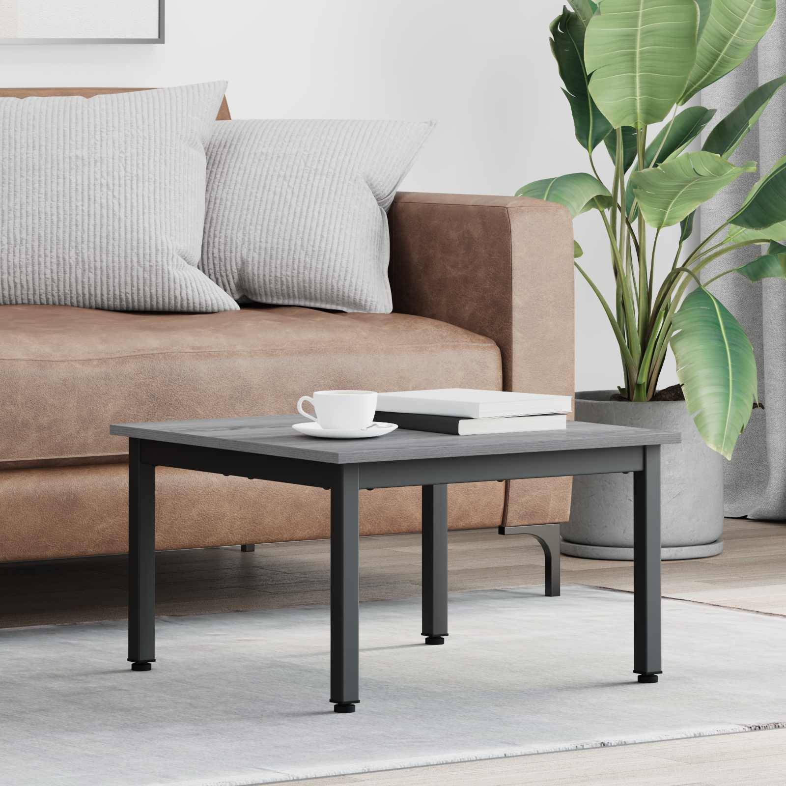 Ein braunes Sofa mit grauen Kissen steht hinter einem schwarzen Couchtisch, auf dem eine weiße Tasse steht. Neben dem Sofa befindet sich eine große grüne Pflanze.