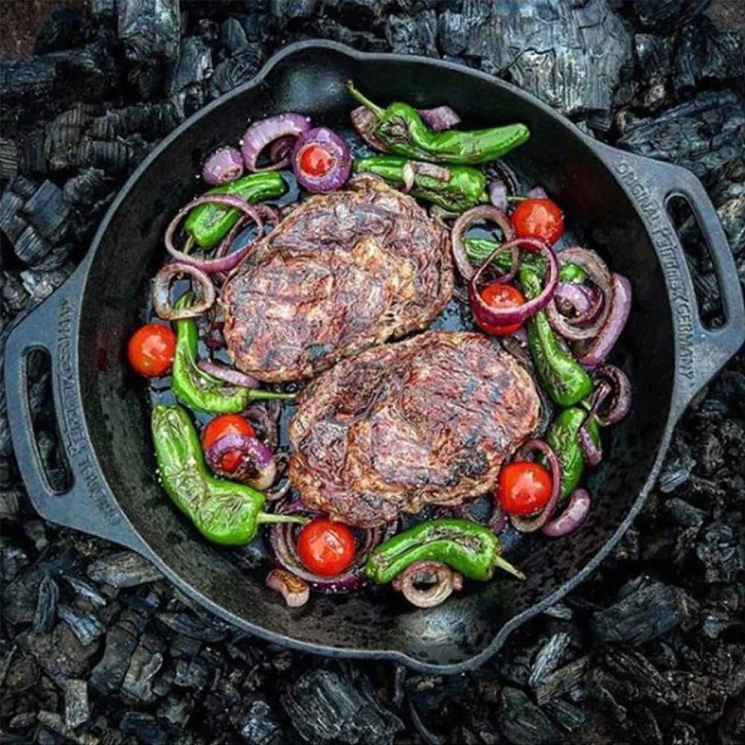 Eine gusseiserne Pfanne mit zwei gegrillten Steaks, gerösteten roten Zwiebeln, grünen Paprikaschoten und Kirschtomaten, die auf verkohlten Kohlen platziert ist.