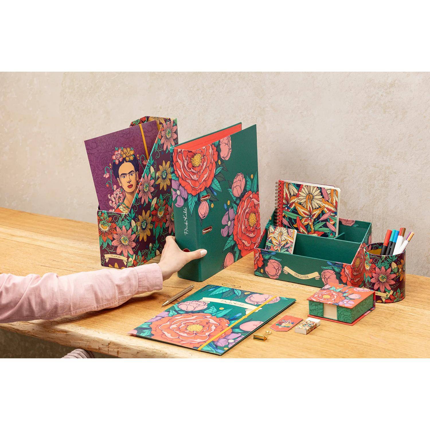 Buntes Schreibwarenset mit Blumen- und künstlerischen Designs auf einem Holzschreibtisch. Enthält Ordner, Boxen, Notizbücher und eine Hand, die auf einen Ordner zugreift.
