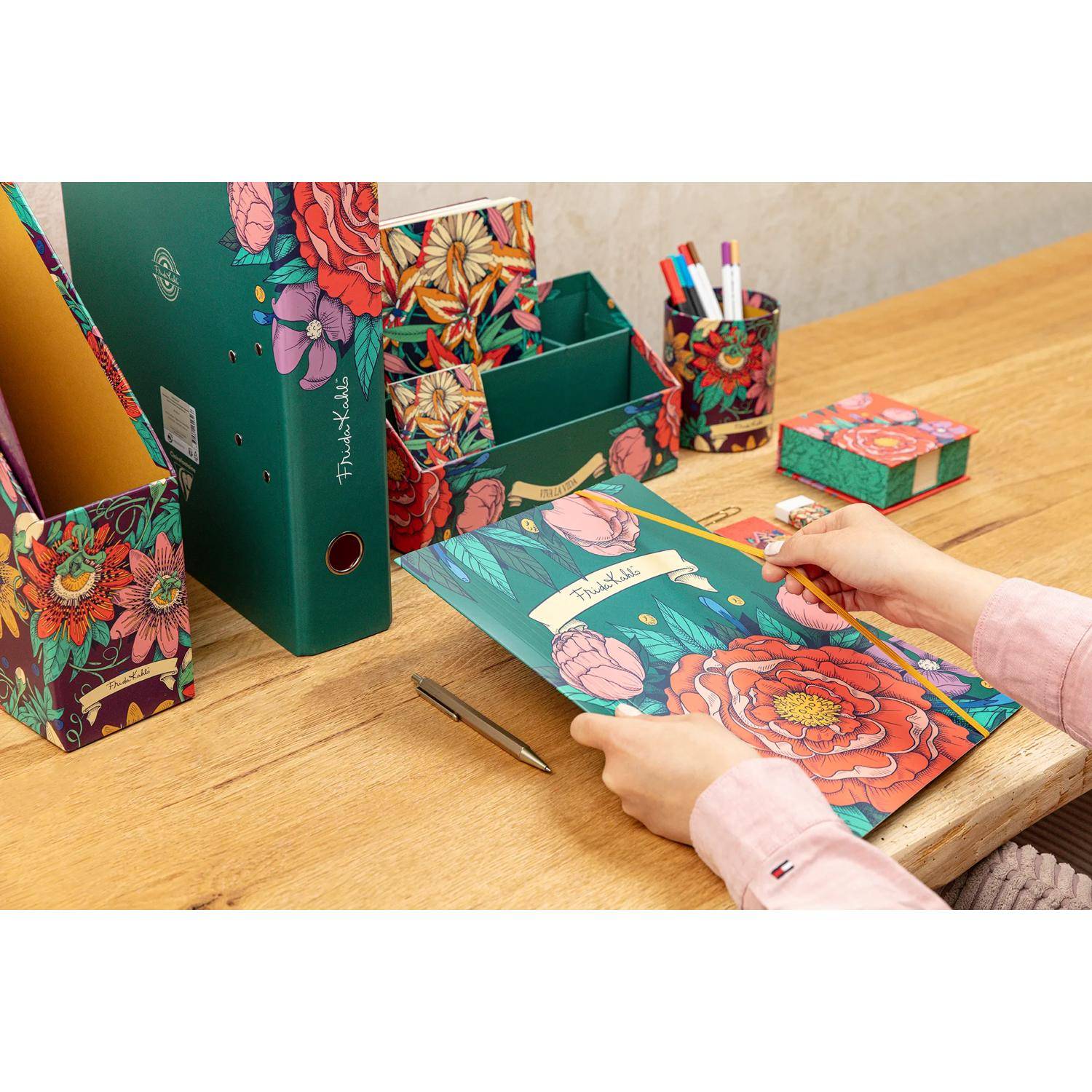 Eine Person arrangiert farbenfrohe, blumenthematische Schreibwaren auf einem Holzschreibtisch, einschließlich eines Ordners, eines Stiftehalters und Dokumentenboxen.