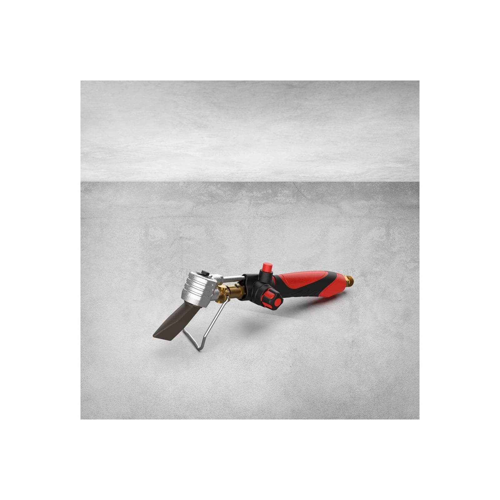 Eine rote und schwarze tragbare Gaslötlampe mit Gasventil, Düse und Metallständer vor einem grauen Betonhintergrund.