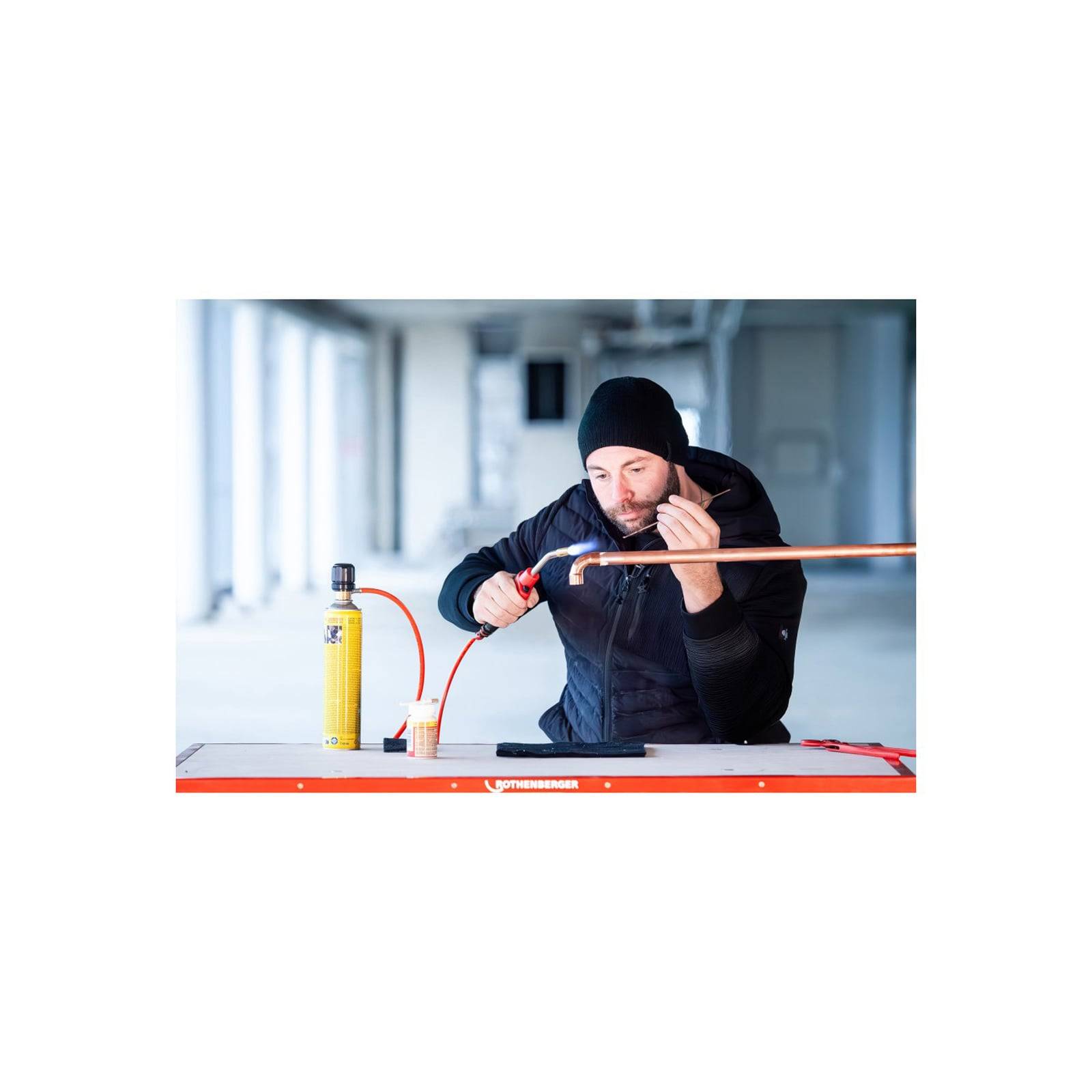 Eine Person mit einer schwarzen Mütze verwendet einen Gasbrenner, um ein Kupferrohr auf einer Werkbank zu löten. Ein gelber Gaszylinder befindet sich in der Nähe.