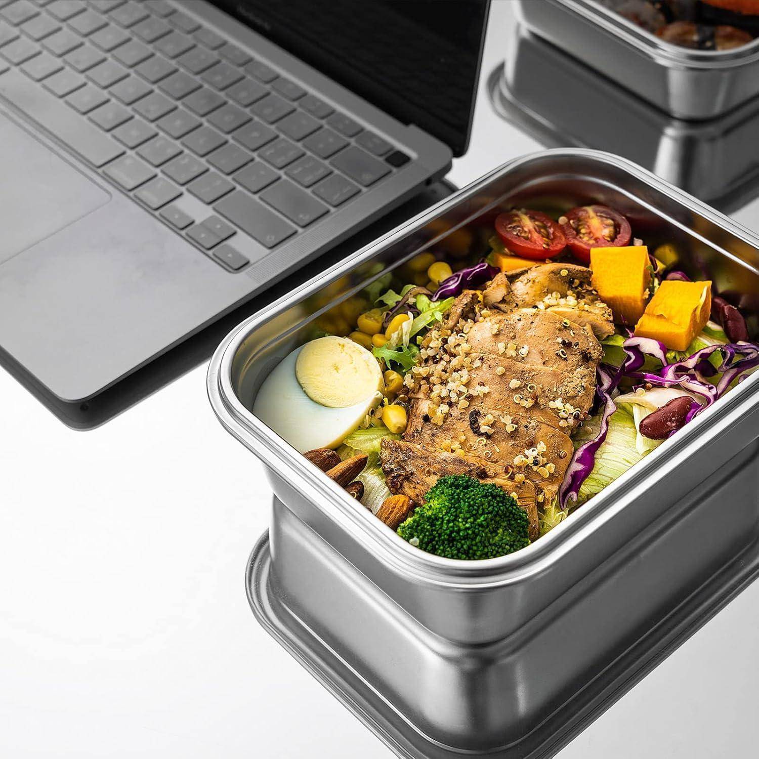 Ein Edelstahlbehälter gefüllt mit einem bunten Salat, der Hähnchen, Kirschtomaten, Brokkoli und Eier enthält, platziert neben einem offenen Laptop.
