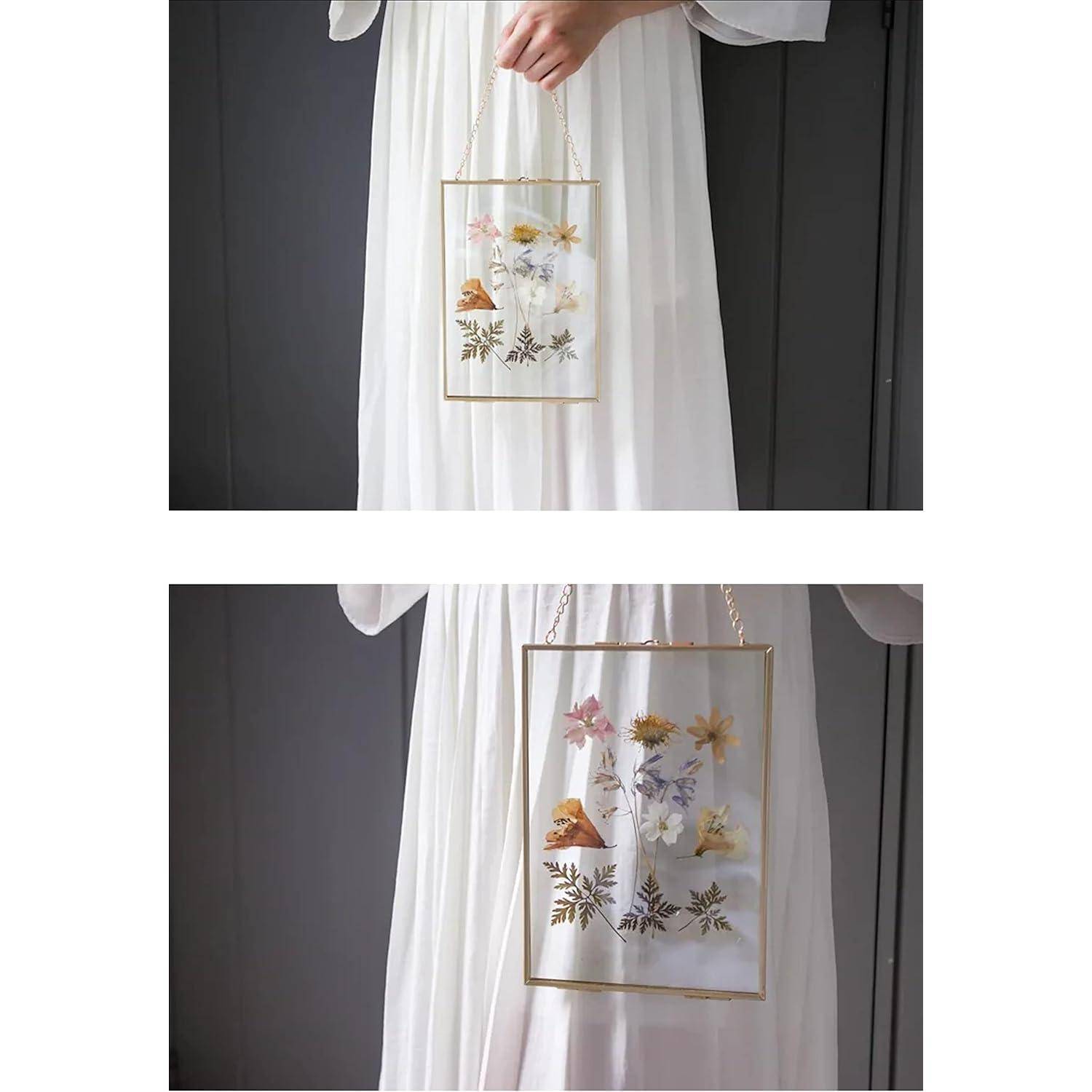 Eine Person in einem weißen Kleid hält einen Glasrahmen mit gepressten Blumen und Blättern und präsentiert das Kunstwerk vor einem dunklen Hintergrund.