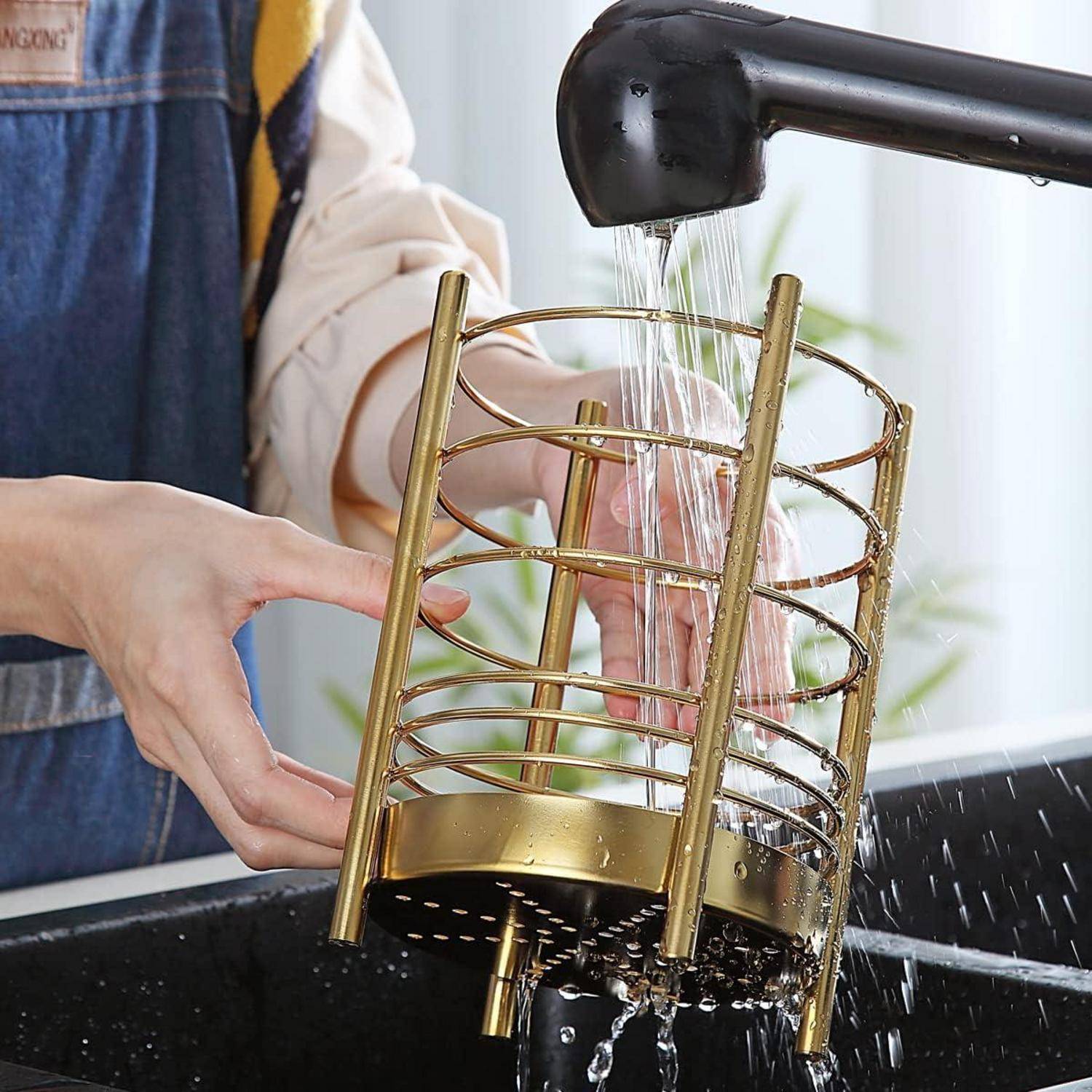 Eine Person, die einen goldfarbenen Drahtkorb unter einem schwarzen Wasserhahn in einem Waschbecken abspült, wobei das Wasser durch die Öffnungen des Korbs fließt.