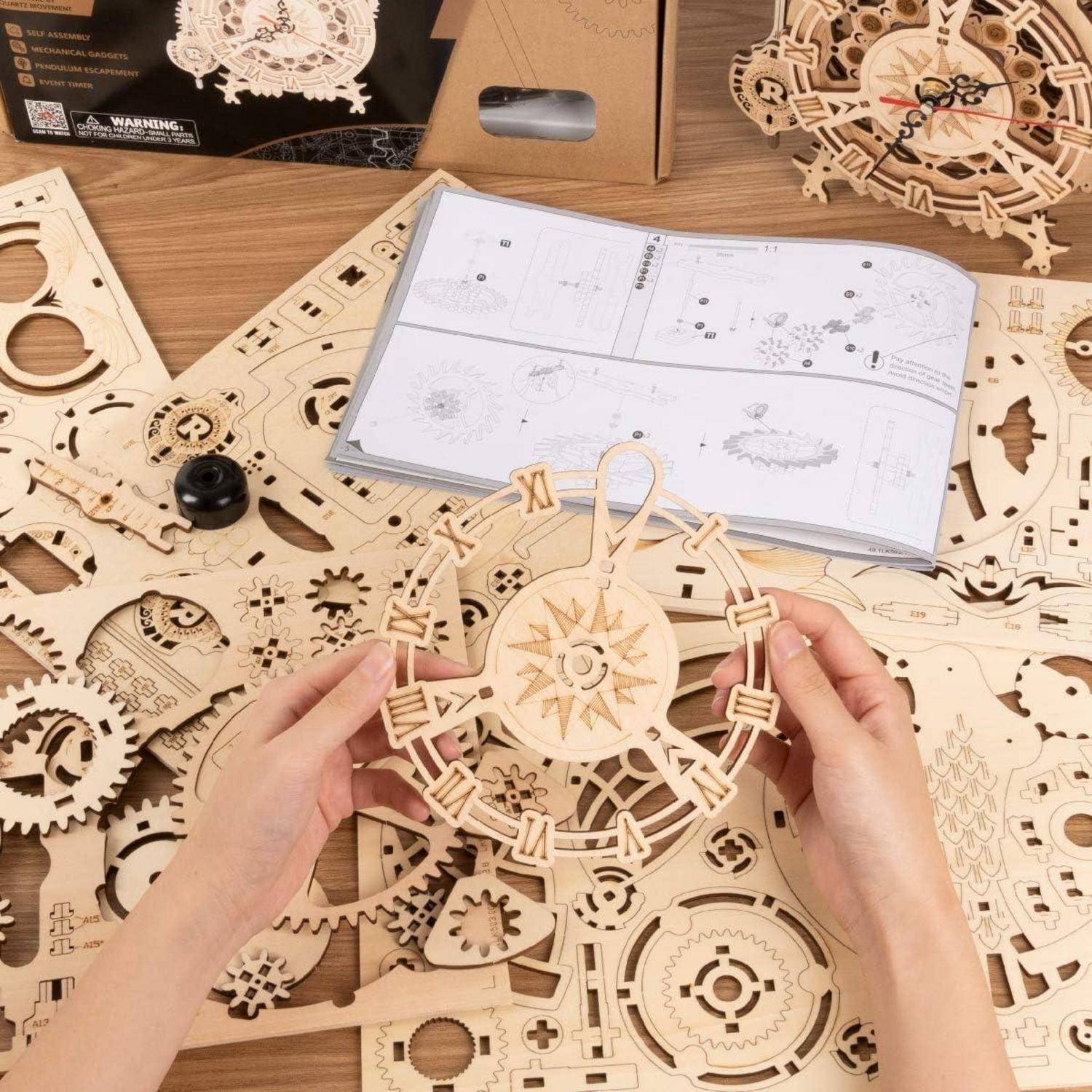 Hände, die ein hölzernes Mechanikmodell mit Zahnrädern zusammenbauen, umgeben von verschiedenen Holzteilen und einem Anleitungsheft auf einem Tisch.