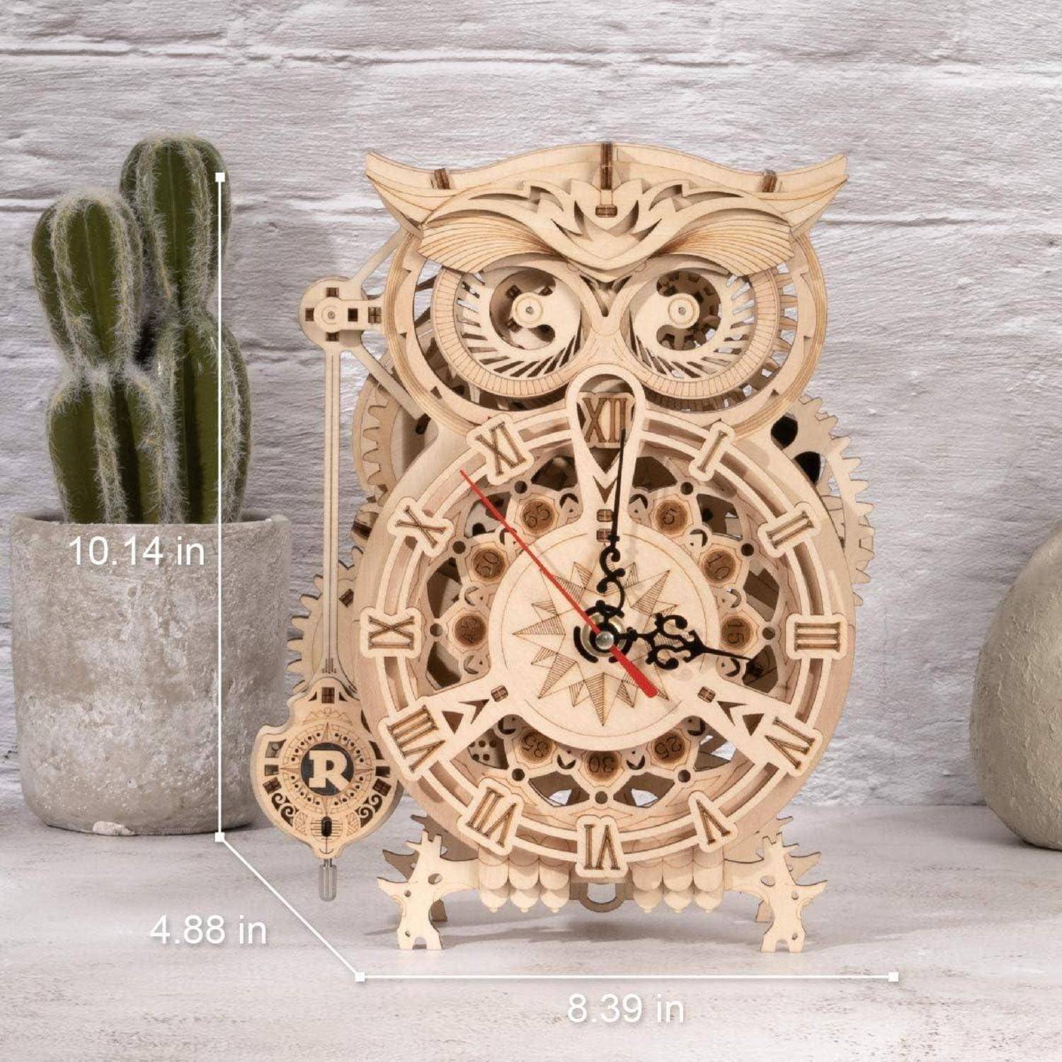Eine hölzerne eulenförmige Uhr mit sichtbaren Zahnrädern steht auf einem Tisch. Die Abmessungen sind markiert: 10,14 in Höhe, 8,39 in Breite, 4,88 in Tiefe. Ein Kaktus befindet sich im Hintergrund.