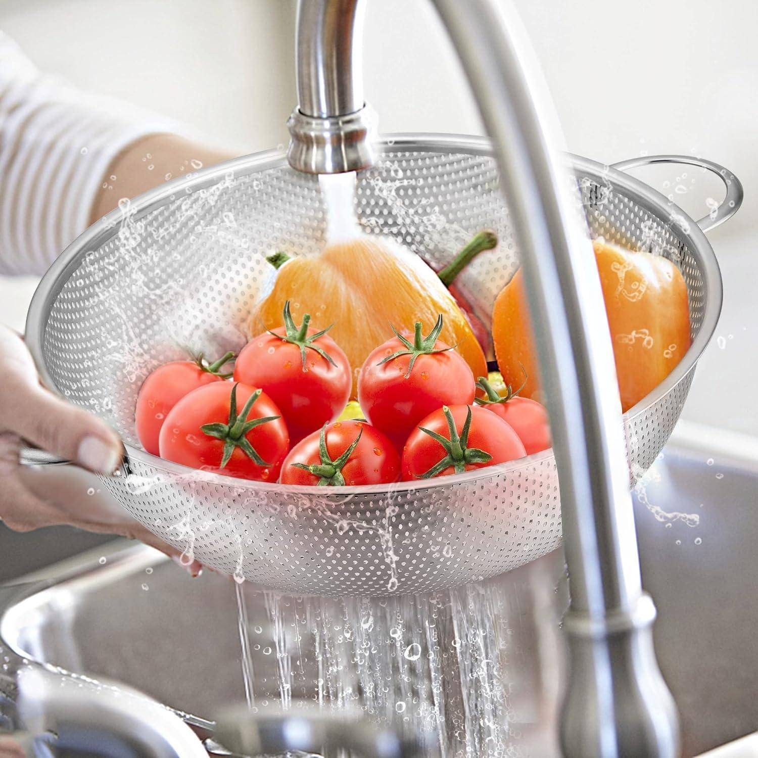 Eine Person wäscht rote und orangefarbene Tomaten in einem Sieb unter fließendem Wasserhahn in einem Küchenbecken, wobei Wassertropfen herumspritzen.
