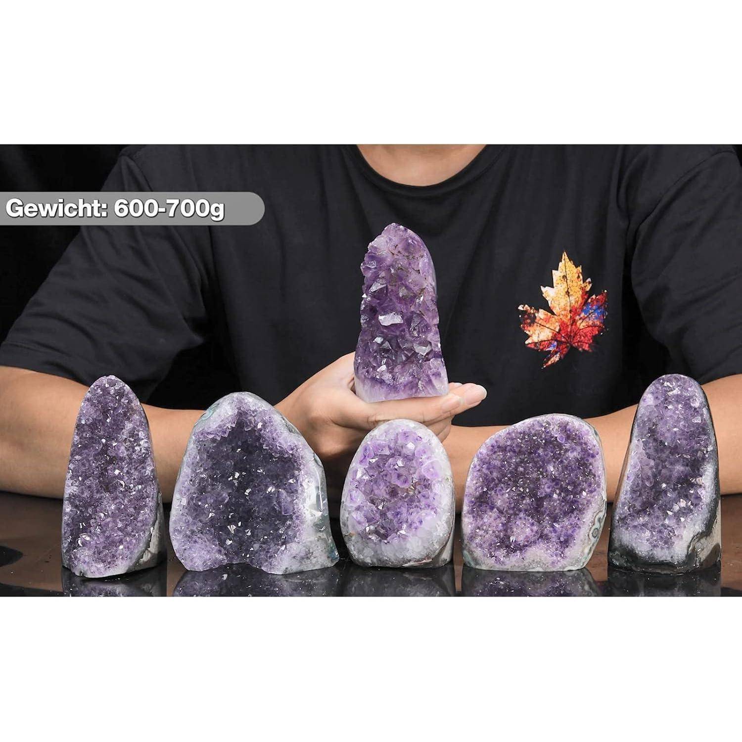 Eine Person hält einen Amethystkristall und präsentiert ihn zusammen mit vier weiteren Kristallen auf einem Tisch. Text lautet: 'Gewicht: 600-700g'.