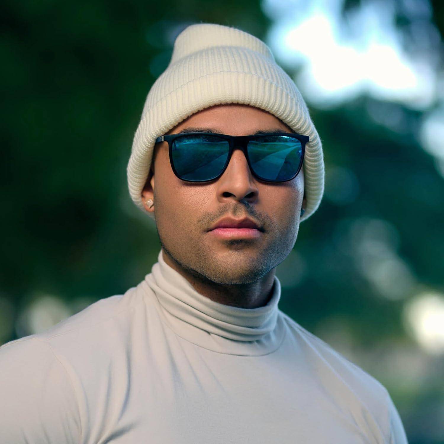 Eine Person mit Sonnenbrille und einer beigen Strickmütze, gekleidet in einen beigen Rollkragenpullover, steht im Freien mit unscharfen grünen Pflanzen im Hintergrund.