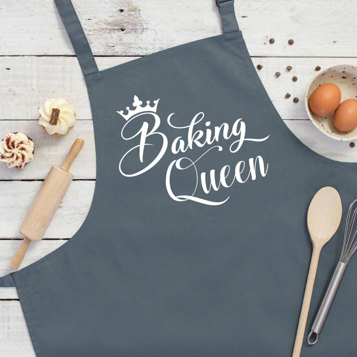 Eine graue Schürze mit den Worten ‚Baking Queen' in weißer Schrift, umgeben von einem Nudelholz, einem Holzlöffel und Eiern auf einer Holzoberfläche.