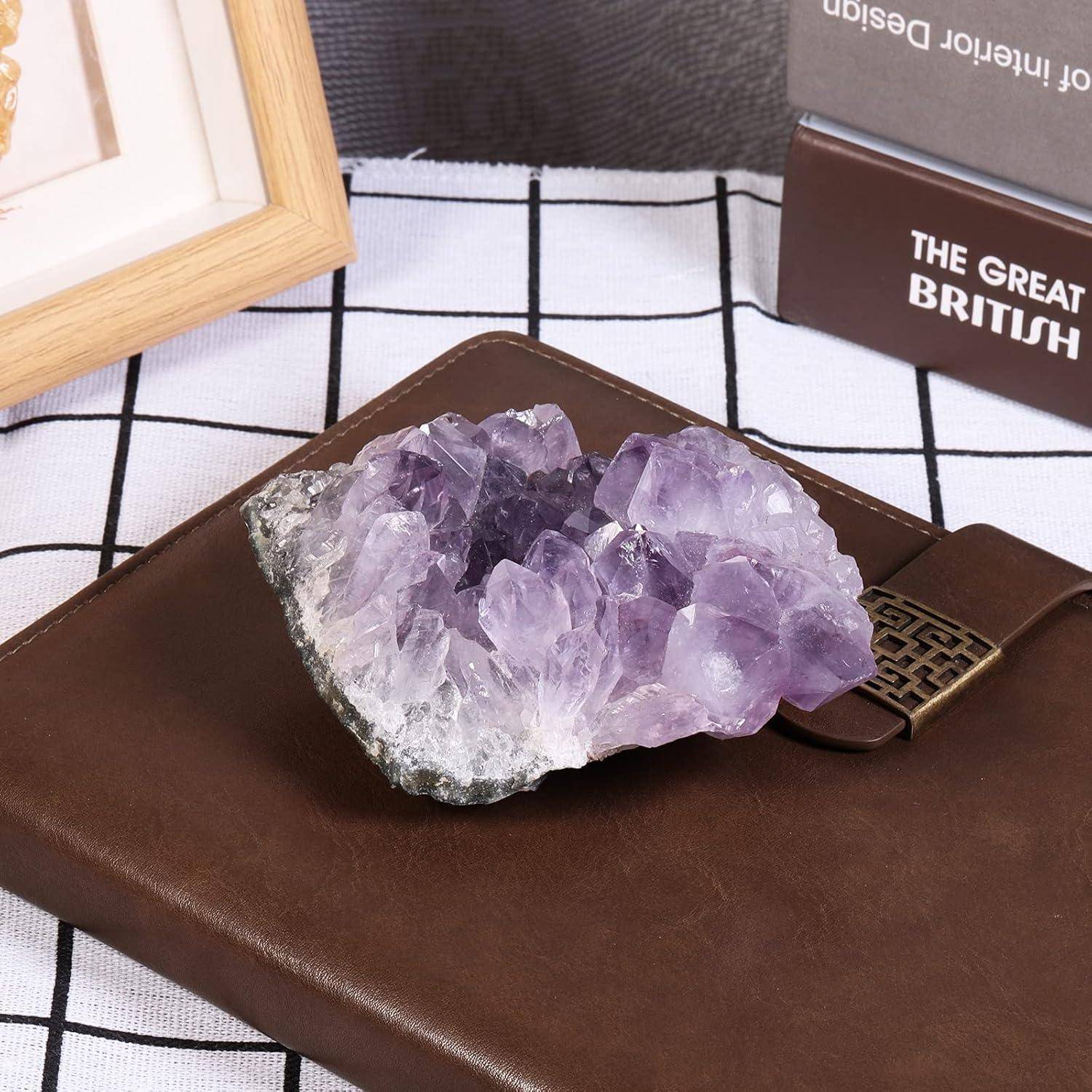 Ein Cluster aus violetten Amethystkristallen ruht auf einem braunen Lederbuch, neben einem Holzrahmen-Bild und einer teilweisen Ansicht eines Buches mit dem Titel 