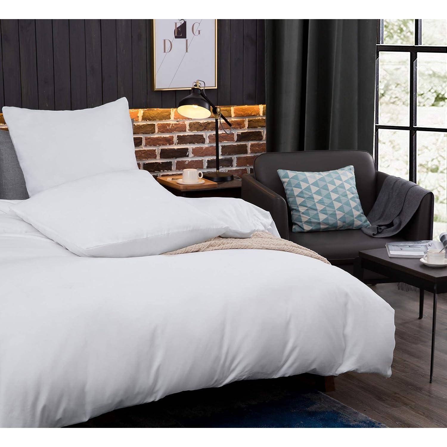 Ein gemütliches Schlafzimmer mit einem ordentlich bezogenen Bett in weißer Bettwäsche, einem weichen grauen Stuhl mit einem geometrischen Kissen und einer Ziegelakzentwand.
