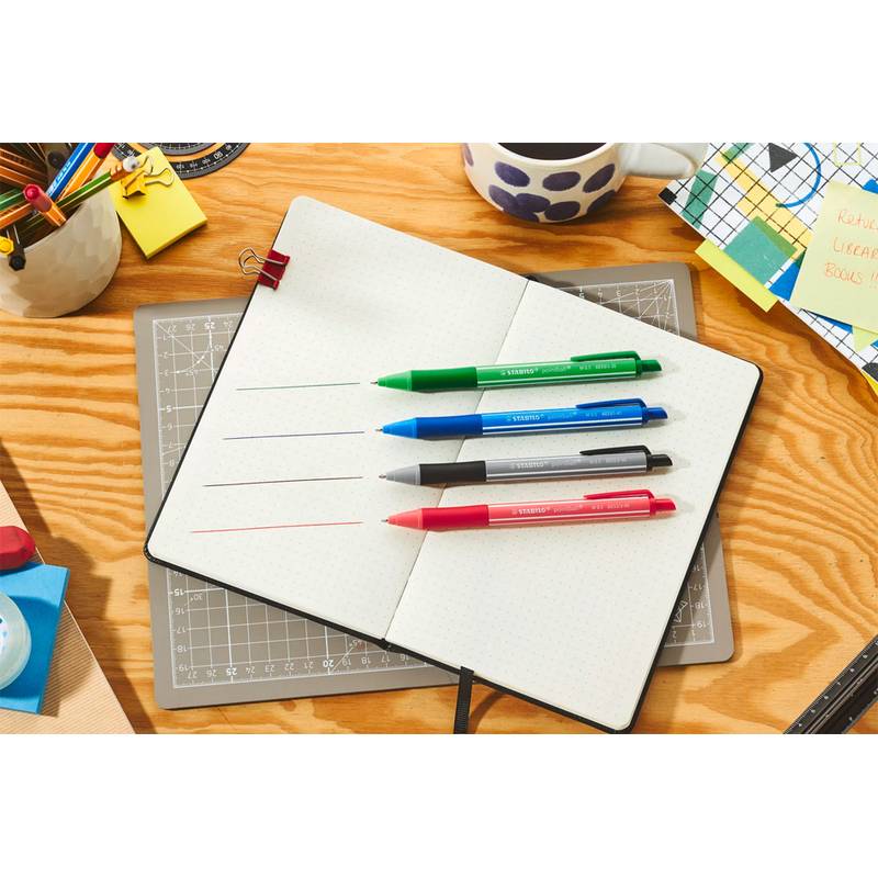 Ein offenes Notizbuch mit Linien, die von grünen, blauen, schwarzen und roten Stiften gezeichnet wurden. Rundherum befinden sich Büromaterialien wie Papiere, ein Lineal und eine Tasse.