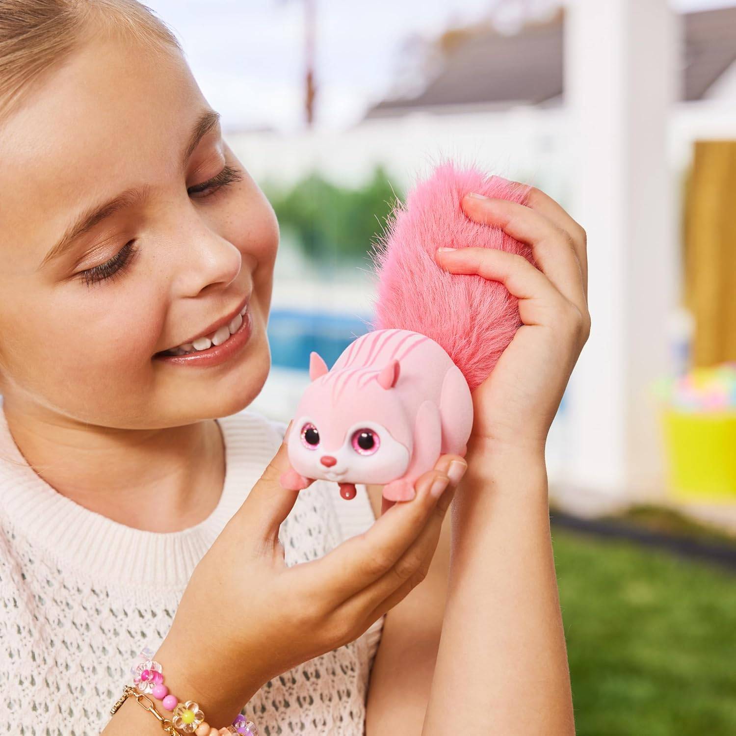 Ein junges Mädchen lächelt, während es ein rosa, flauschig-geschwänztes Spielzeug hält, das einem Eichhörnchen ähnelt, im Freien mit bunter Spielausrüstung im Hintergrund.