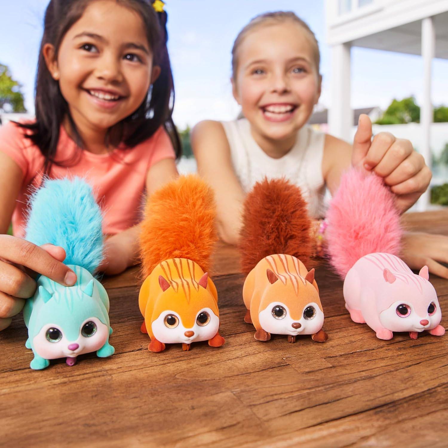 Zwei lächelnde Kinder präsentieren bunte Spielzeugkreaturen mit flauschigen Schwänzen auf einer Holzoberfläche im Freien.