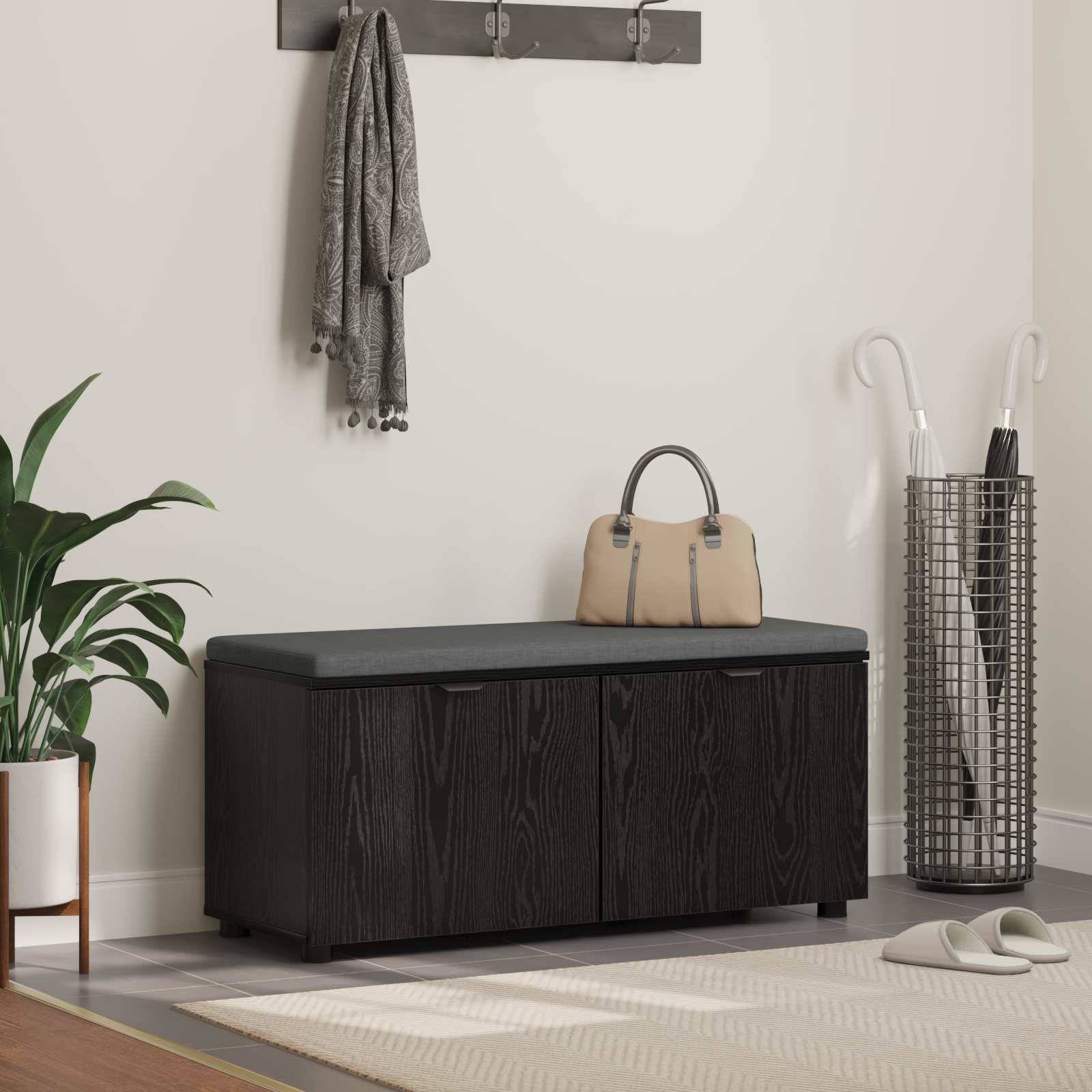 Eine Flurszene mit einer schwarzen Holzlagerbank mit grauem Kissen. Darüber hängen zwei graue Schals an einer Wandgarderobe. Pflanzen schmücken den Raum.