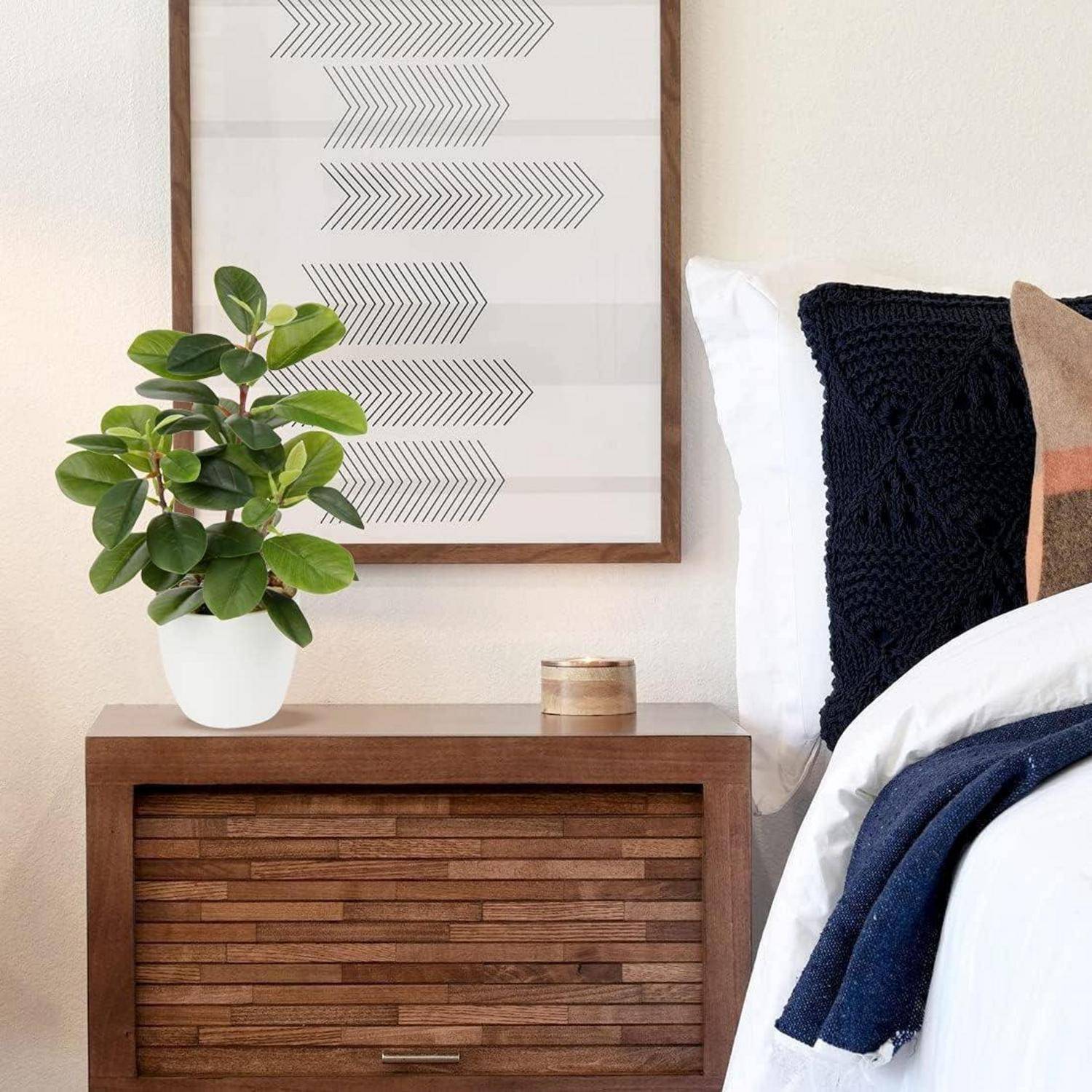 Eine minimalistische Schlafzimmerecke mit einem Holznachttisch, auf dem eine Topfpflanze und eine Kerze stehen, neben einem ordentlich gemachten Bett mit einem dunklen Dekokissen.