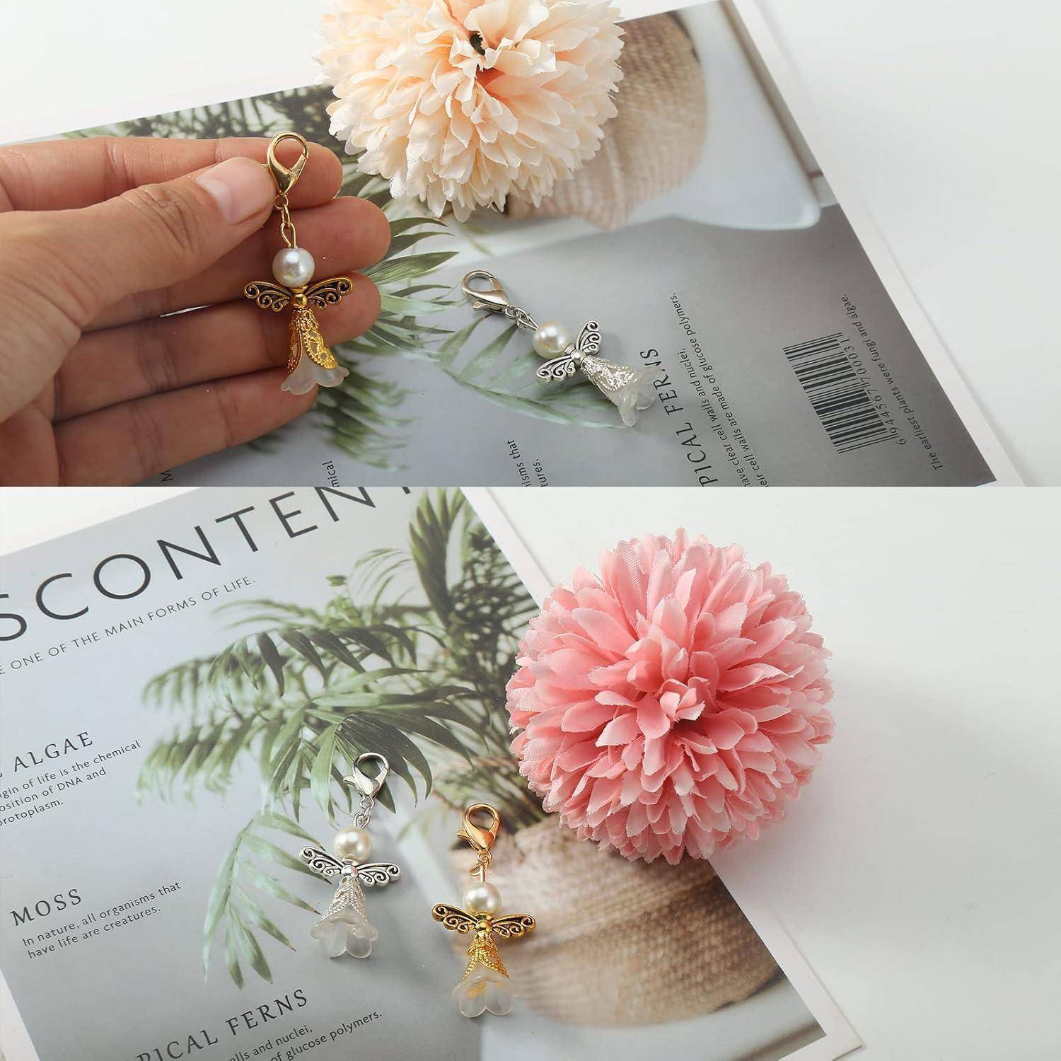 Eine Hand, die einen engelsförmigen Schlüsselanhänger mit einer Perle hält. Darunter sind zwei ähnliche Schlüsselanhänger auf einer Zeitschrift mit einer rosa Blume platziert.