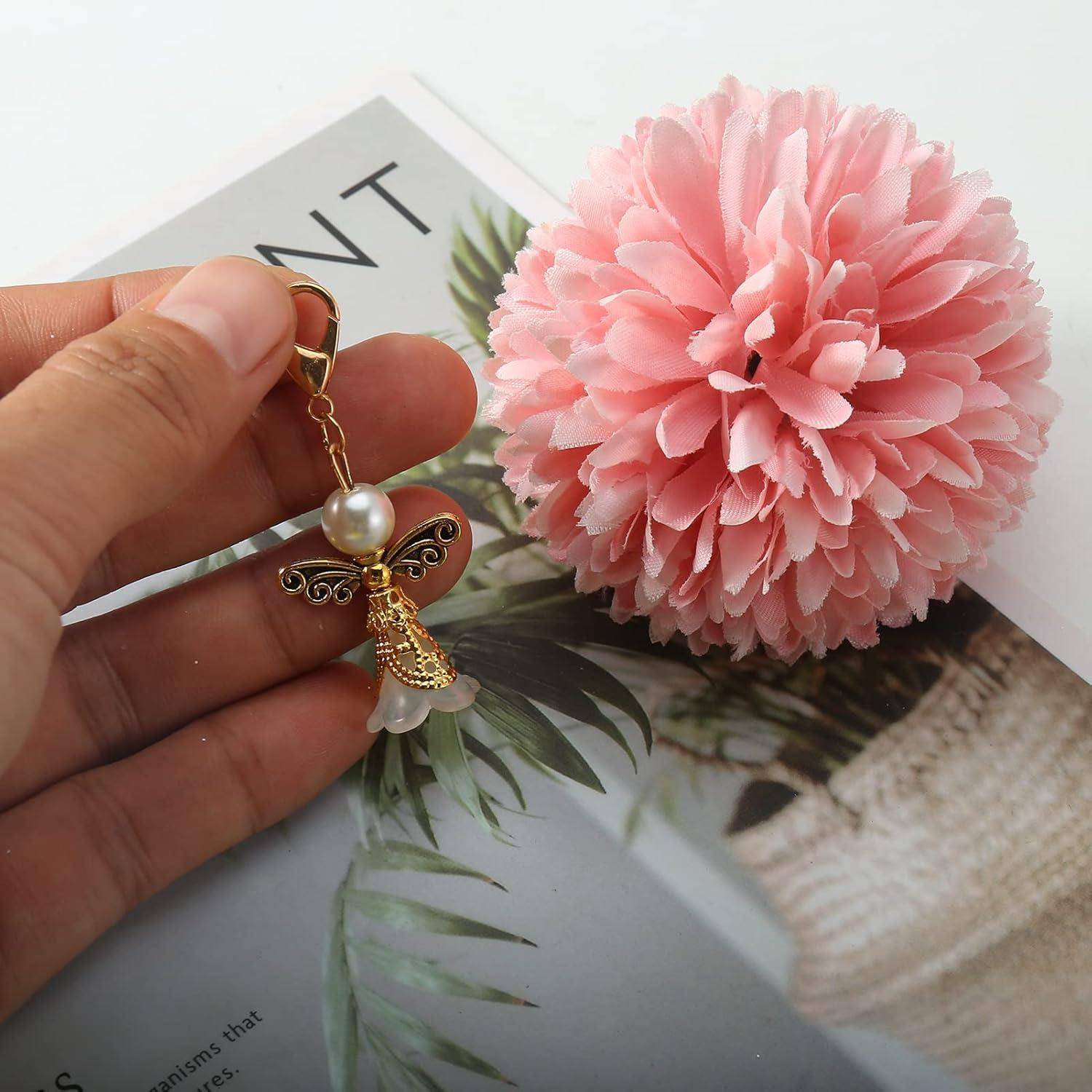 Eine Hand hält einen dekorativen Engelanhänger mit Perle und goldenen Details, neben einer rosa Pompom-Blume auf einer Magazinseite.