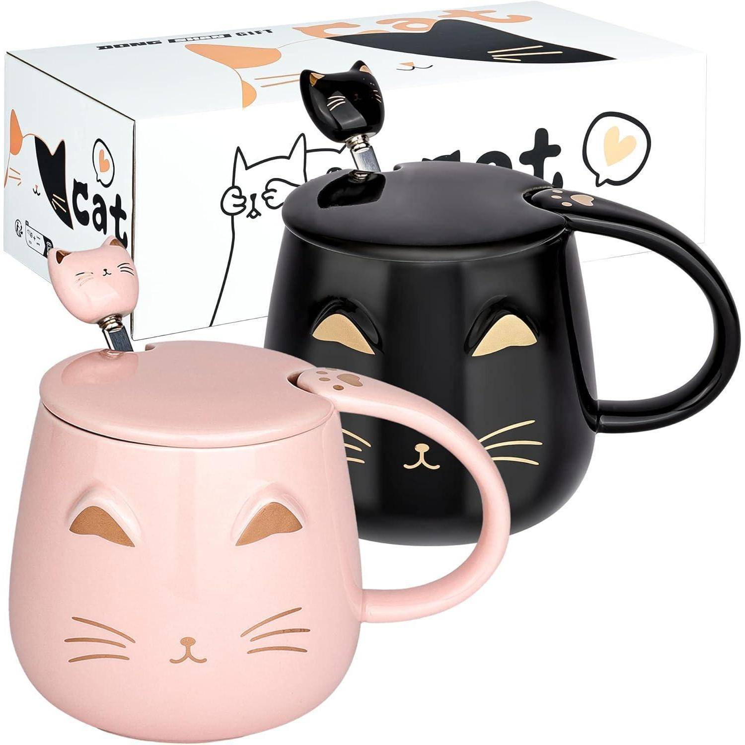 Zwei Keramikbecher mit Katzengesichtern, einer in Rosa und einer in Schwarz, jeweils mit passendem Deckel und Löffel, wobei die Henkel eine kleine Katzenfigur aufweisen.